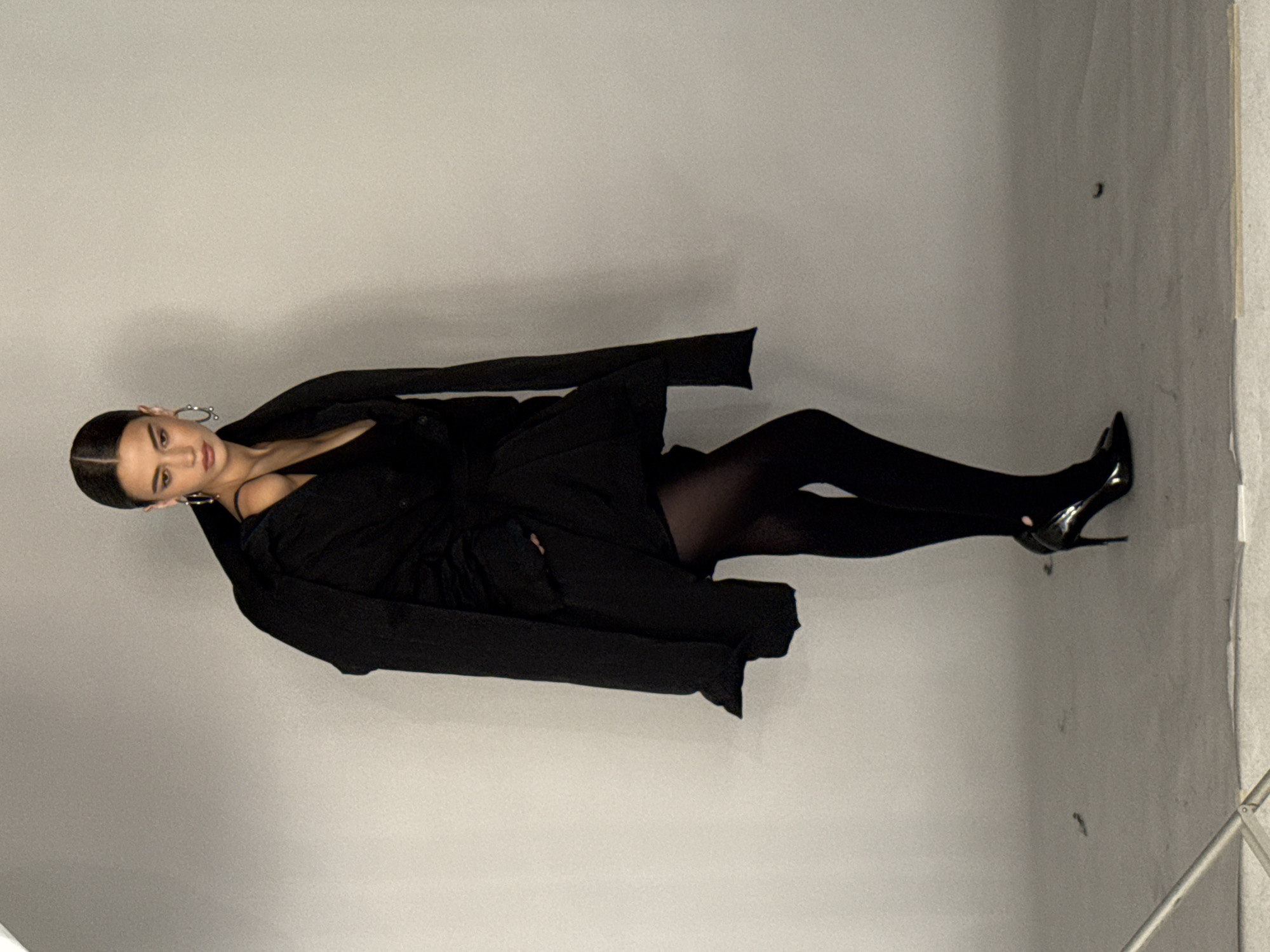 A woman stands against a plain gray background in a studio setting. She is wearing an oversized black jacket draped over a black dress, black pantyhose, and black high heels. Her hair is pulled back in a bun, and she is wearing large hoop earrings. She is looking towards the camera with a neutral expression. The lighting is soft and diffused, creating a subtle shadow on the background. The floor appears to be a light gray concrete.