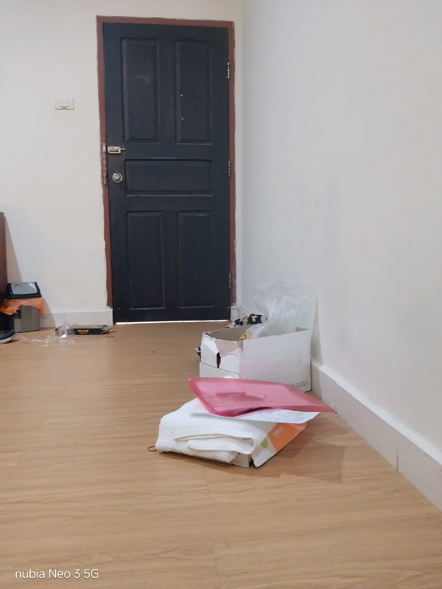 The image depicts a domestic indoor scene, likely a hallway or entryway, with a dark wooden door in the background. The floor is made of light-colored wood laminate. To the left of the door, a light-colored wall features a white electrical outlet with two visible sockets. In the foreground, several items are placed on the floor: a stack of white folded items, possibly towels or linens, topped with a bright pink folder or binder. Beside these items, an open cardboard box is visible, containing some crumpled white plastic and what appears to be fabric. To the far left, a dark trash can with an orange liner is partially visible. Further to the left and closer to the door, scattered plastic packaging and a black box are also present on the floor. The lighting suggests a bright, possibly daytime, indoor environment. The lower left corner of the image displays the text "nubia Neo 3 5G", indicating the device used to capture the photograph. The location of Tembilahan, Indonesia, is context provided separately and not visually discernible from the image itself.