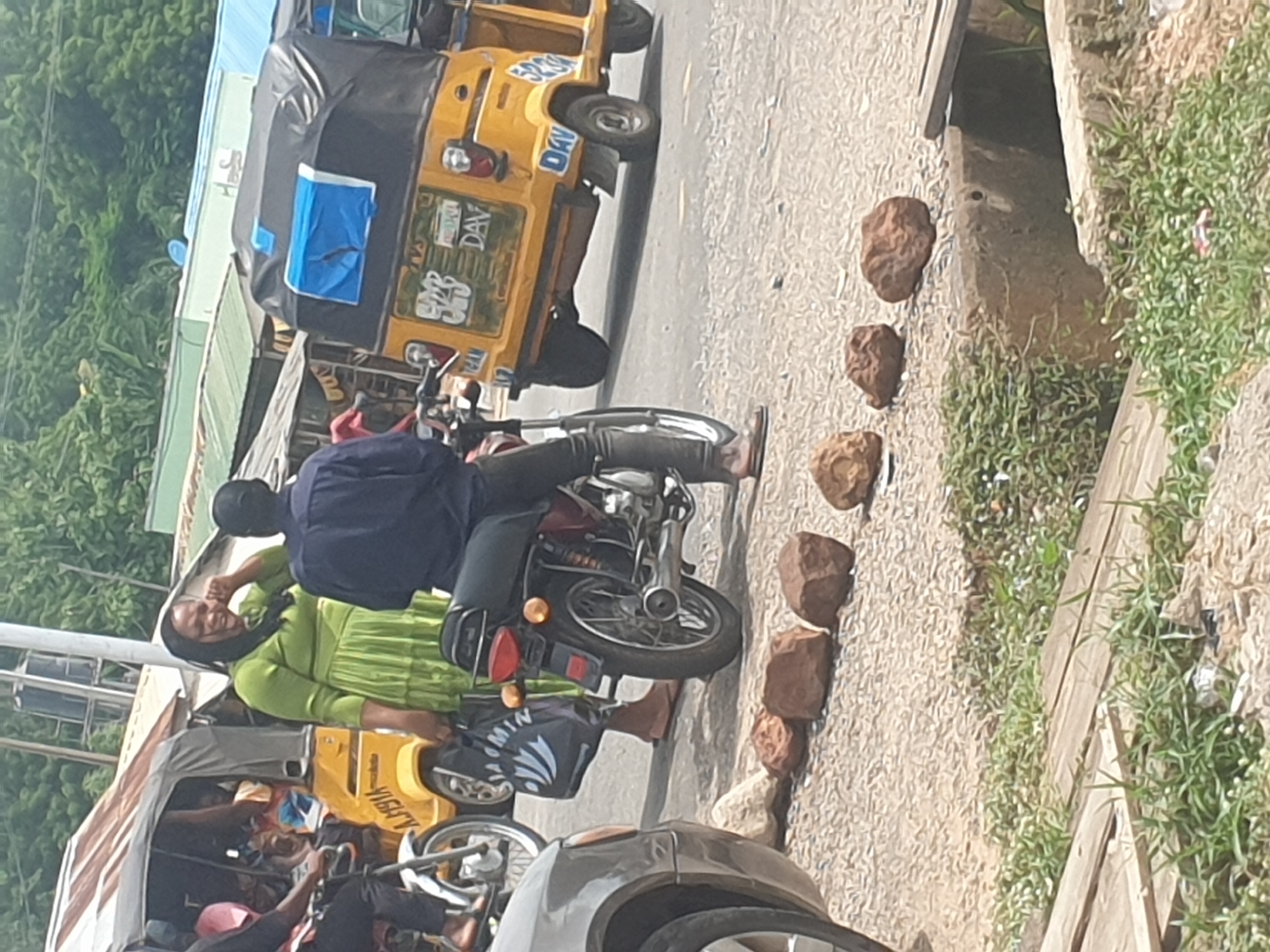 A street in an urban area is shown, with a man on a motorcycle wearing a dark blue shirt and black pants. He is barefoot and appears to be preparing to start the motorcycle. Next to him, a woman in a green dress is reaching out to touch his shoulder, and she seems to be smiling. In the background, there are yellow motorized tricycles, known as "keke napeps," common in some African cities. The street is paved, and a line of rocks is placed near the edge of the road next to a patch of grass. The vegetation is lush, and various other vehicles are present on the street, indicating a busy traffic environment.