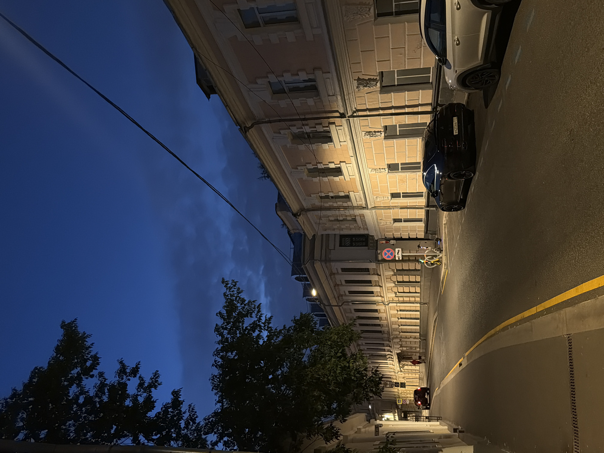 An exterior, eye-level, wide shot shows a street in Moscow, Russia, at dusk or early evening. The sky is a deep, dark blue with some clouds. A tree with dark green leaves is on the left side of the frame, casting a shadow. Buildings with pale yellow facades and white decorative trim line the right side of the street. They have multiple windows and arched doorways. Streetlights illuminate the road.
On the street, there are two parked cars: a black sedan and a white SUV. A yellow line divides the road. A person is walking a dog on the sidewalk in the distance. A bicycle and a scooter are parked on the sidewalk near a traffic sign. Another car is visible driving away in the distance on the left. The scene conveys a sense of quiet urban life at the end of the day.