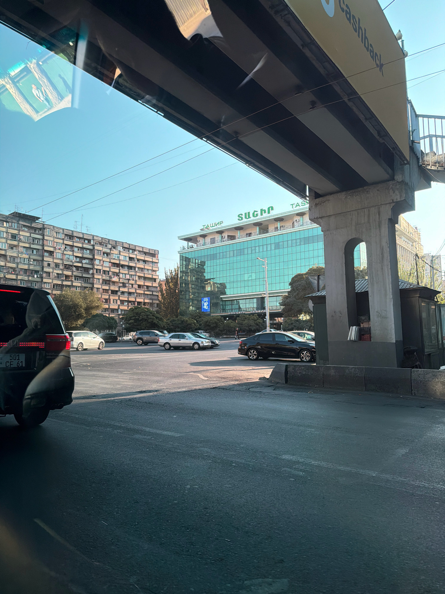 The image shows a street scene in Yerevan, Armenia, with a prominent overpass casting a shadow over the road. In the background, a large, modern building with a teal-colored glass facade is visible, bearing the Cyrillic inscription "ТАШИР". To the left of this building is a tall, multi-story apartment complex with numerous balconies. Several cars are on the road, some in motion and others appearing to be stopped or waiting. The sky is clear and blue, suggesting daylight. The foreground shows the asphalt road surface and a section of the overpass support structure.