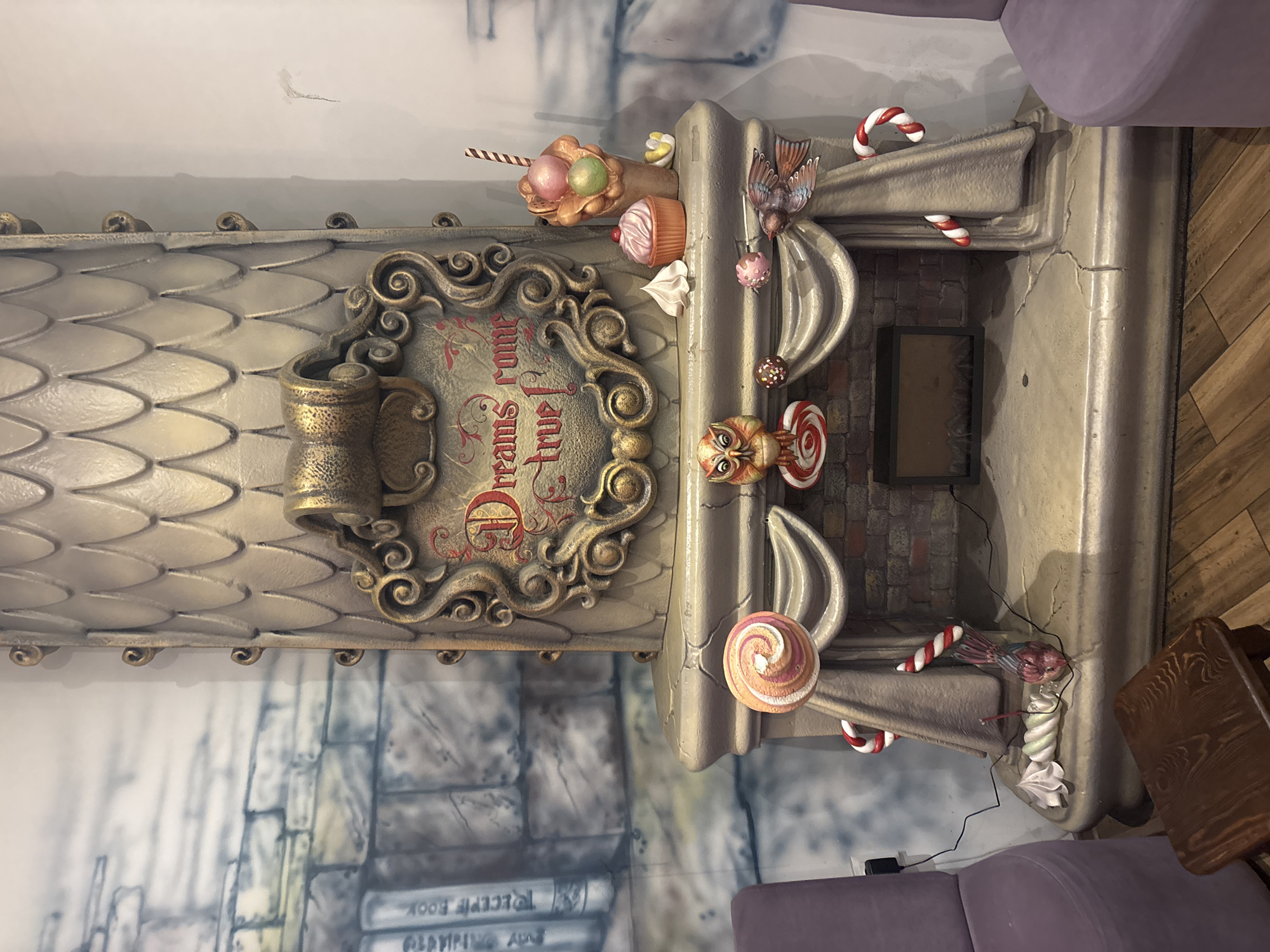 The scene depicts an interior space, likely a commercial establishment, in the city of Unknown, Unknown, featuring a highly decorative, whimsical fireplace structure. The central subject is a large, light-gold colored fireplace, approximately 2.5 meters in height. Its upper section, resembling a chimney breast, is covered in a scalloped or scale-like texture and tapers upward. Centered on the upper half is an ornate, gold-framed plaque with the stylized red text "Dreams come true!". The mantlepiece below is adorned with various dessert and candy-themed sculptures. On the left are a large, swirl lollipop and an owl figure with a spiral lollipop base. On the right are a waffle cone sundae with multiple scoops, a pink cupcake with a cherry, a white meringue, and a yellow candy. A bird sculpture with spread wings is positioned on the right side of the mantle. Sculpted fabric swags hang below the mantle, featuring red and white candy cane ornaments. The firebox area is brick-textured with a dark, rectangular insert. The base of the fireplace shows faux cracks. To the left of the fireplace, a purple upholstered armchair is partially visible, with a black power adapter plugged into a wall outlet behind it. A small, dark brown wooden stool sits on the wooden floor in front of the armchair. On the floor near the left side of the fireplace, a broken candy cane and a twisted meringue-like sculpture are present, with a black wire trailing nearby. To the right, another purple armchair is partially visible. The wall to the left of the fireplace features a mural depicting a grey stone wall with several book spines, including one labeled "RECIPE BOOK". The wall to the right is a plain, light color. The floor is covered with light brown wooden planks laid in a diagonal pattern. No individuals are present.