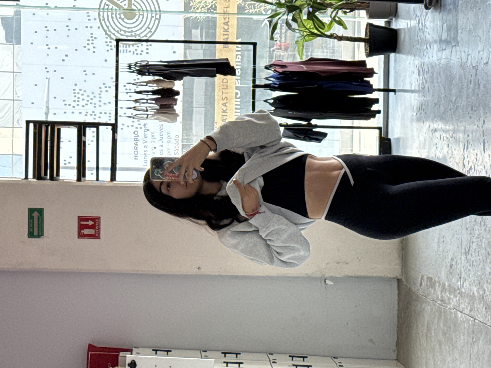 A young woman is captured taking a mirror selfie indoors, in what appears to be a retail store or studio in Puebla, Mexico. The image is rotated 90 degrees clockwise. She holds her phone up to her face, looking into the screen, with her long dark hair falling over her shoulder. She is dressed in athletic wear, including a light grey cropped hoodie, a black sports bra with white trim, and black leggings with similar white detailing.
The background reveals a spacious, well-lit interior, predominantly illuminated by natural light streaming through large windows. Clothing items are displayed on black racks behind her, suggesting a commercial establishment. On the window, a visible schedule reads "HORARIO" (Schedule), indicating operating hours: "Lunes a Viernes" (Monday to Friday) from "am - 2 pm" and "pm - 9 pm," and "Sábado" (Saturday) until "2 pm." Other inverted text visible on the window includes fragments like "RIKASTUD" and "EAIKASFLUC," possibly parts of a business name. A potted plant sits on a sill or shelf towards the right.
On a light-colored wall, two signs are present: a green sign with an upward arrow and a red sign featuring a fire extinguisher symbol. The inverted text on the red sign likely says "EXTINTOR" (Fire Extinguisher). The floor is a light, textured surface, possibly concrete or large tiles. The scene conveys a casual moment captured within a functional, brightly lit commercial space during the daytime.