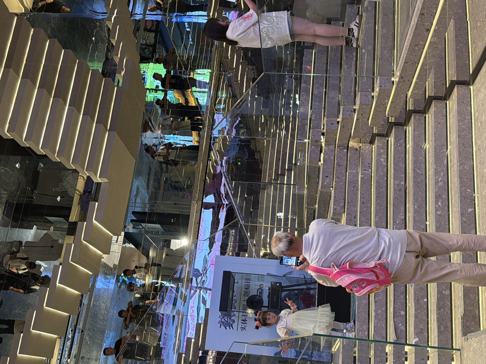 In Yuzhong District, China, an outdoor public space features a multi-level staircase with mirrored walls and ceilings, creating a disorienting visual effect. A young girl, dressed in a light yellow dress and adorned with pink hair accessories, poses with her hands in a peace sign, positioned on a lower landing. An older man, wearing a plaid shirt and beige pants, stands behind her, holding a smartphone to take her photograph. He is carrying a bright pink backpack. Further up the stairs, a woman in a white t-shirt and denim shorts is visible, also holding a smartphone. The mirrored surfaces reflect numerous individuals and the illuminated staircase structure, adding to the complex visual environment.