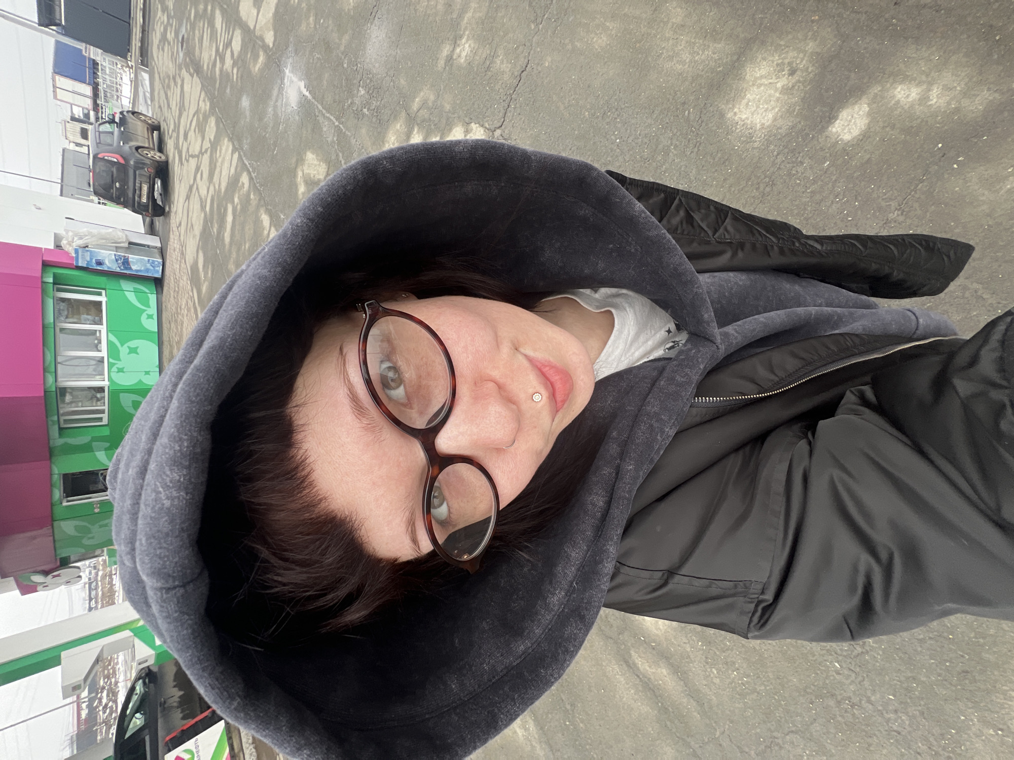 A person wearing glasses, a gray hoodie, and a black jacket is taking a selfie. The location appears to be an outdoor area in the city of Kamennoye, Russia, based on the distinct architecture. A small black car is parked near a green and pink building that has an image of flowers. The ground is gray, possibly concrete.