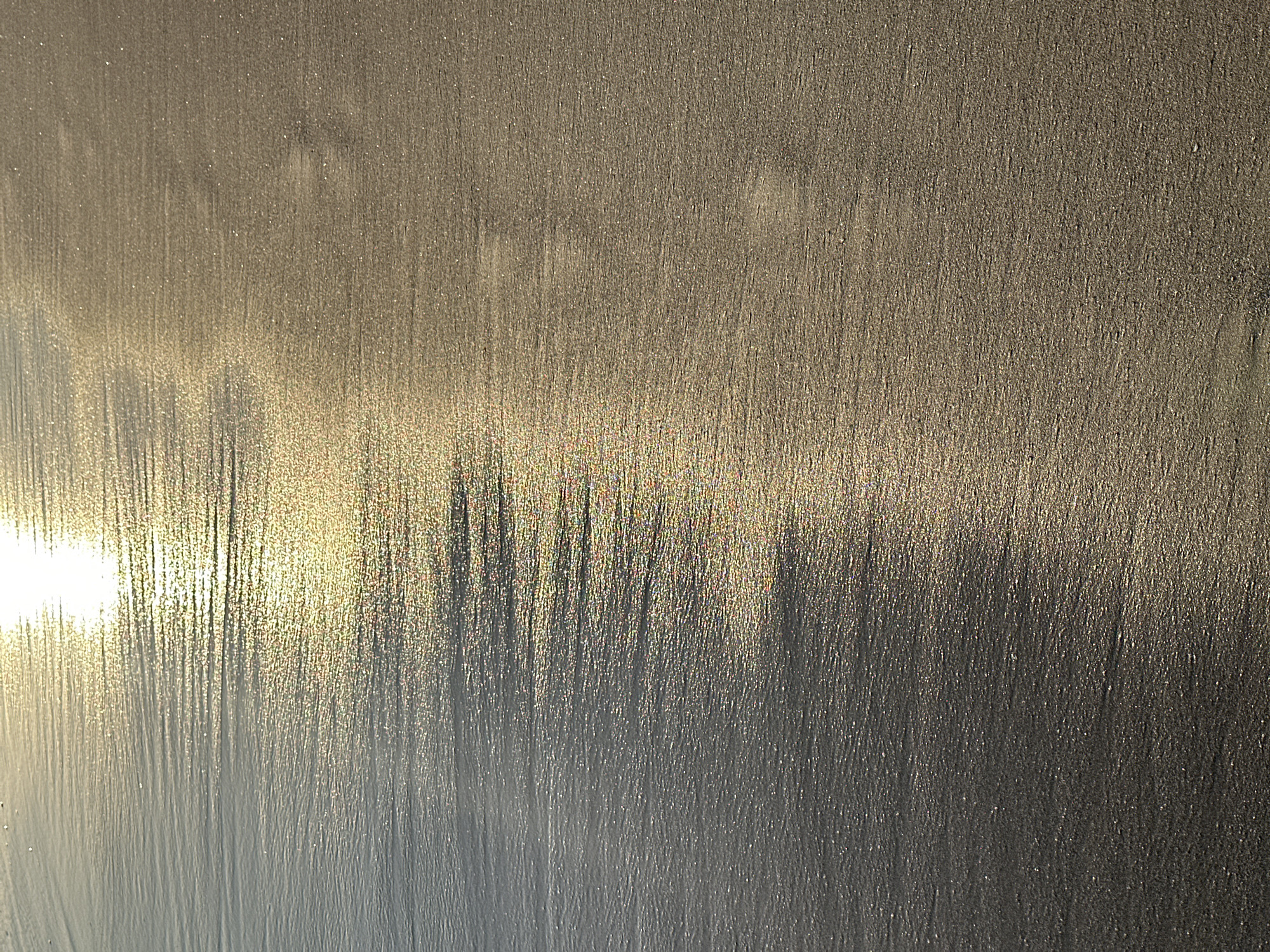 The image displays a close-up, abstract view of wet sand with streaks of light. The texture of the sand appears fine and granular, with visible lines or ripples running vertically, suggesting the movement of water or wind. The lighting creates a strong contrast, with a bright, luminous area on the left side of the frame, likely caused by the sun reflecting off the wet surface. This bright area transitions into darker, shadowed streaks extending towards the right.
The overall impression is that of a natural, outdoor scene, possibly a beach at a particular time of day when the light is low and intense. The location context of North Kuta, Indonesia, implies a tropical beach environment. The play of light and shadow on the sand could indicate either sunrise or sunset, given the golden hues and the long, vertical streaks. There are no people, objects, or visible text in the image. The focus is purely on the texture and light interaction with the sand, evoking a sense of calm and natural beauty. The granular sparkle visible in some areas hints at the presence of tiny shell fragments or mica within the sand.