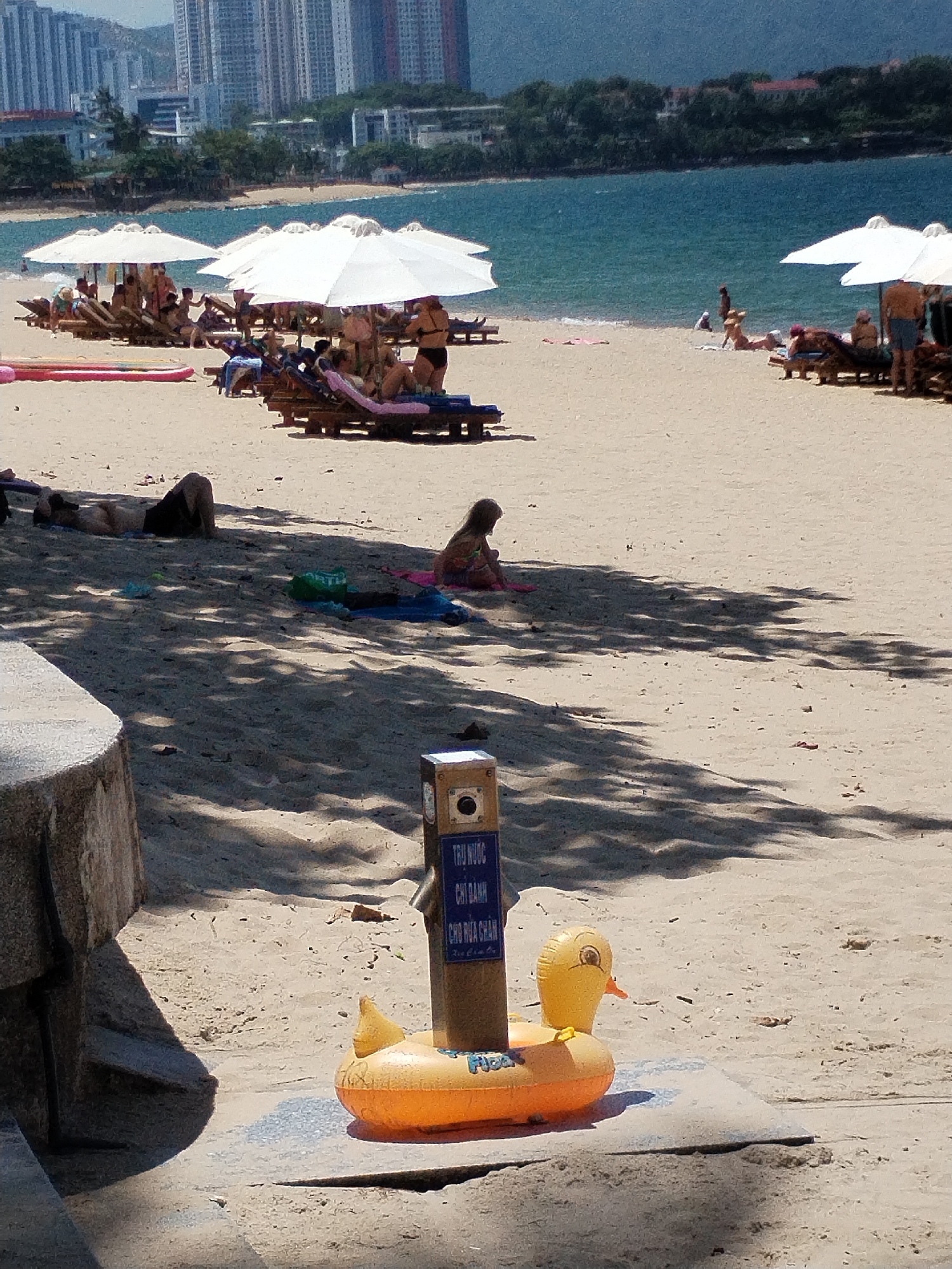 The image depicts a sunny day on a crowded beach in Nha Trang Ward, Vietnam. In the foreground, an orange inflatable duck-shaped float is partially submerged in the sand. Behind it, a brown cylindrical post with a blue sign containing Vietnamese text is visible, with a visible camera lens or sensor. The beach is populated with numerous sunbathers lounging on wooden beach chairs under large white umbrellas. People are scattered across the sand, some lying down, others sitting or walking. The sea is calm and blue in the background, with a cityscape of tall buildings and trees lining the coast. Shadows from palm trees create a dappled effect on the sand.