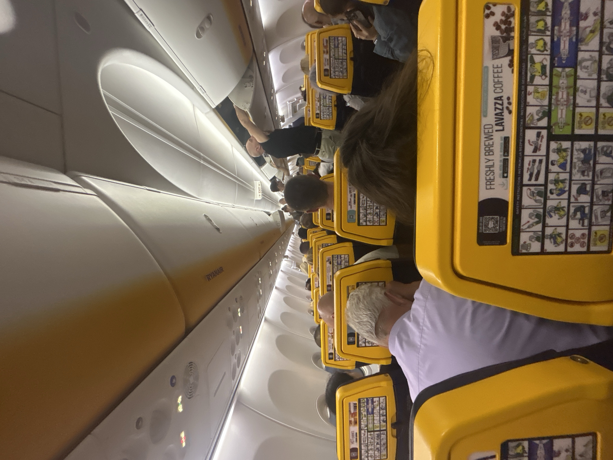 The image depicts the interior of a Ryanair aircraft, likely during a flight. Passengers are seated in rows with yellow headrests, each featuring safety instructions and advertisements for Lavazza coffee. One passenger is standing in the aisle, reaching towards an overhead compartment. The Ryanair logo is visible on the overhead panel. The location of Luqa, Malta, cannot be determined from this image.