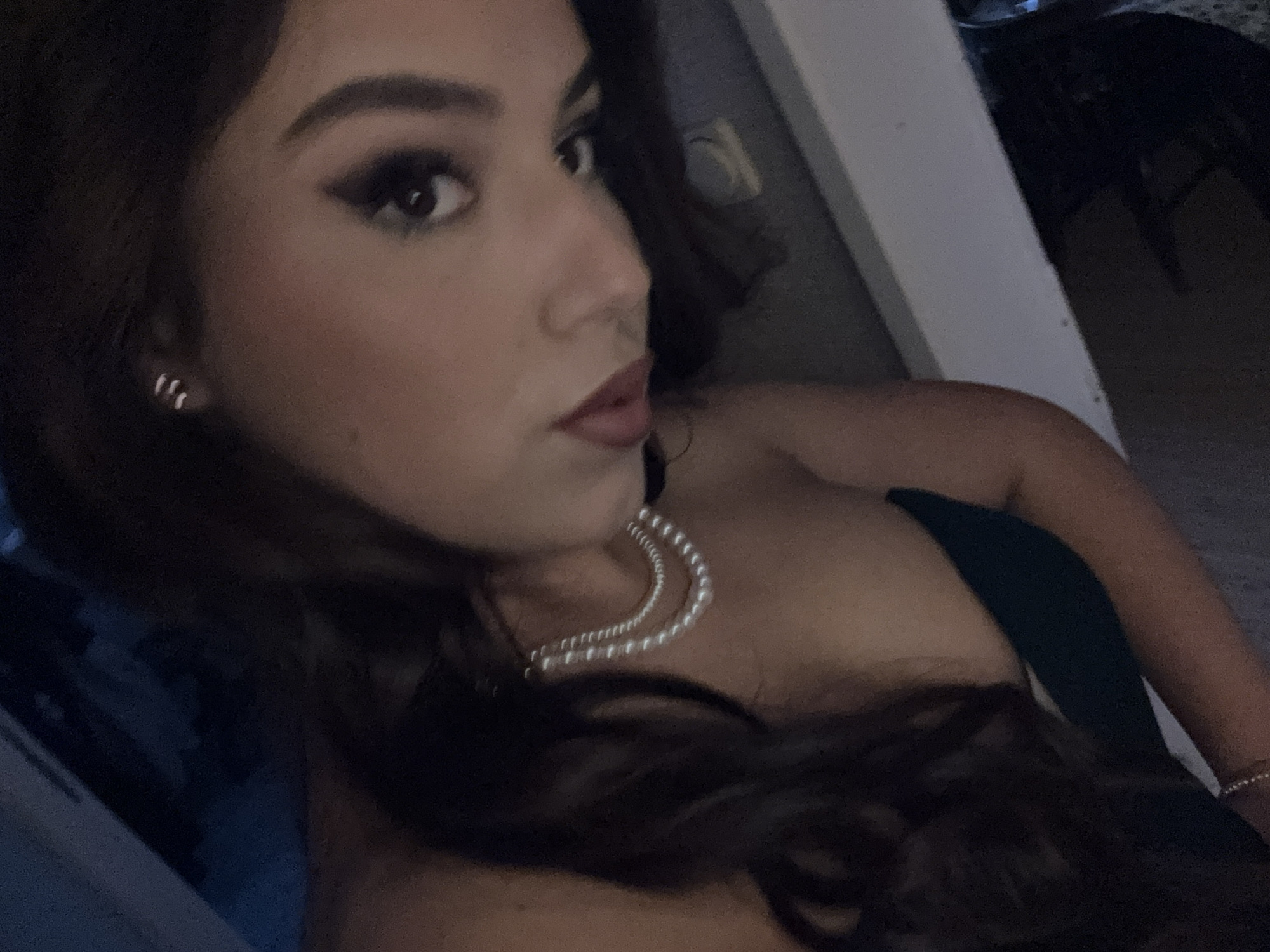 A close-up shot features a woman with dark hair, facing left, wearing a pearl necklace and a dark dress. Her makeup includes a defined eye look and dark lipstick. She has multiple earrings in her left ear. The background is blurred, showing a white or light-colored object and a dark chair. Location: Unknown, Unknown.