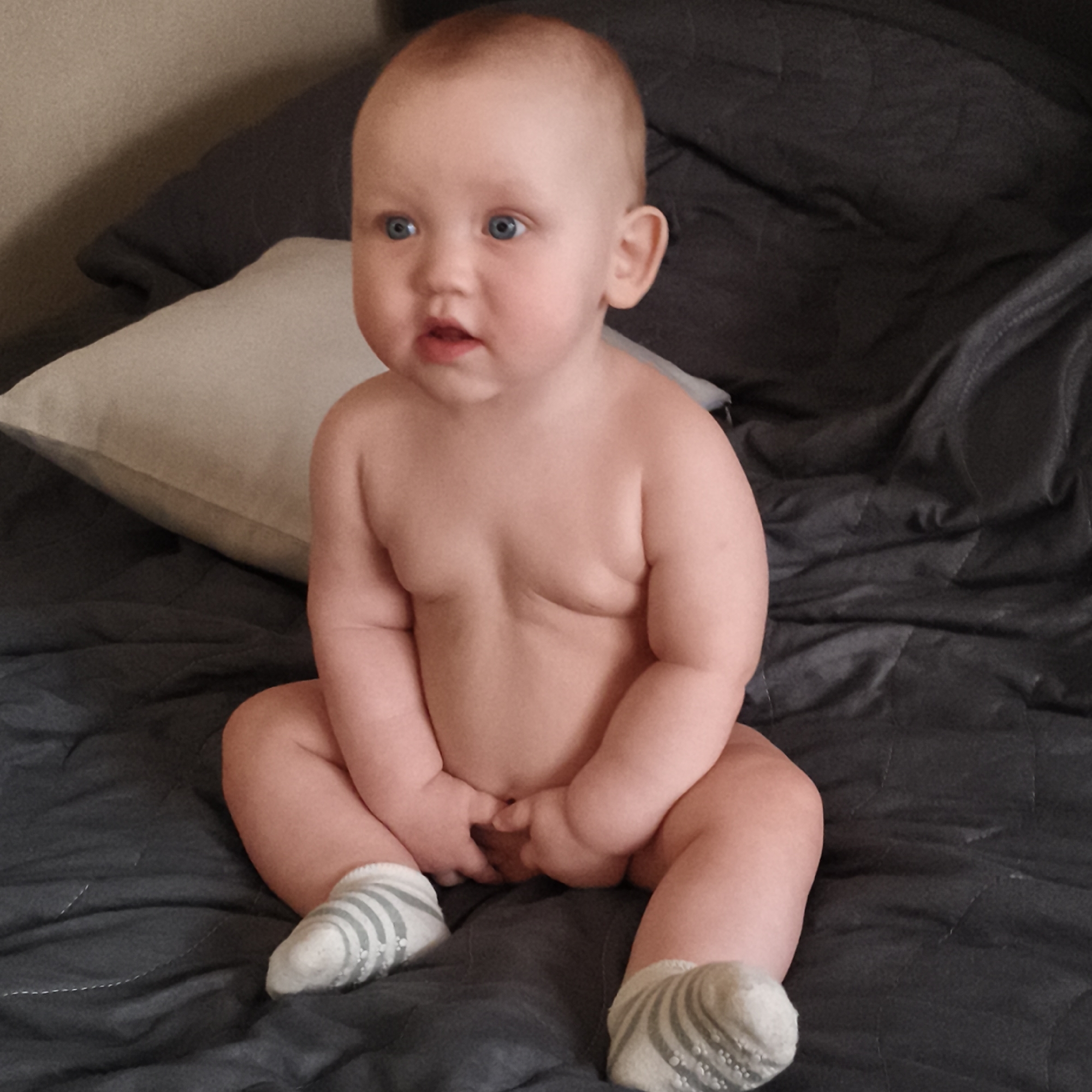 A baby sits on a gray quilted blanket wearing gray and white striped socks. The baby is fair-skinned with blue eyes, looking off to the left. Behind the baby is a light gray pillow and a gray blanket. The location is Unknown, Unknown.