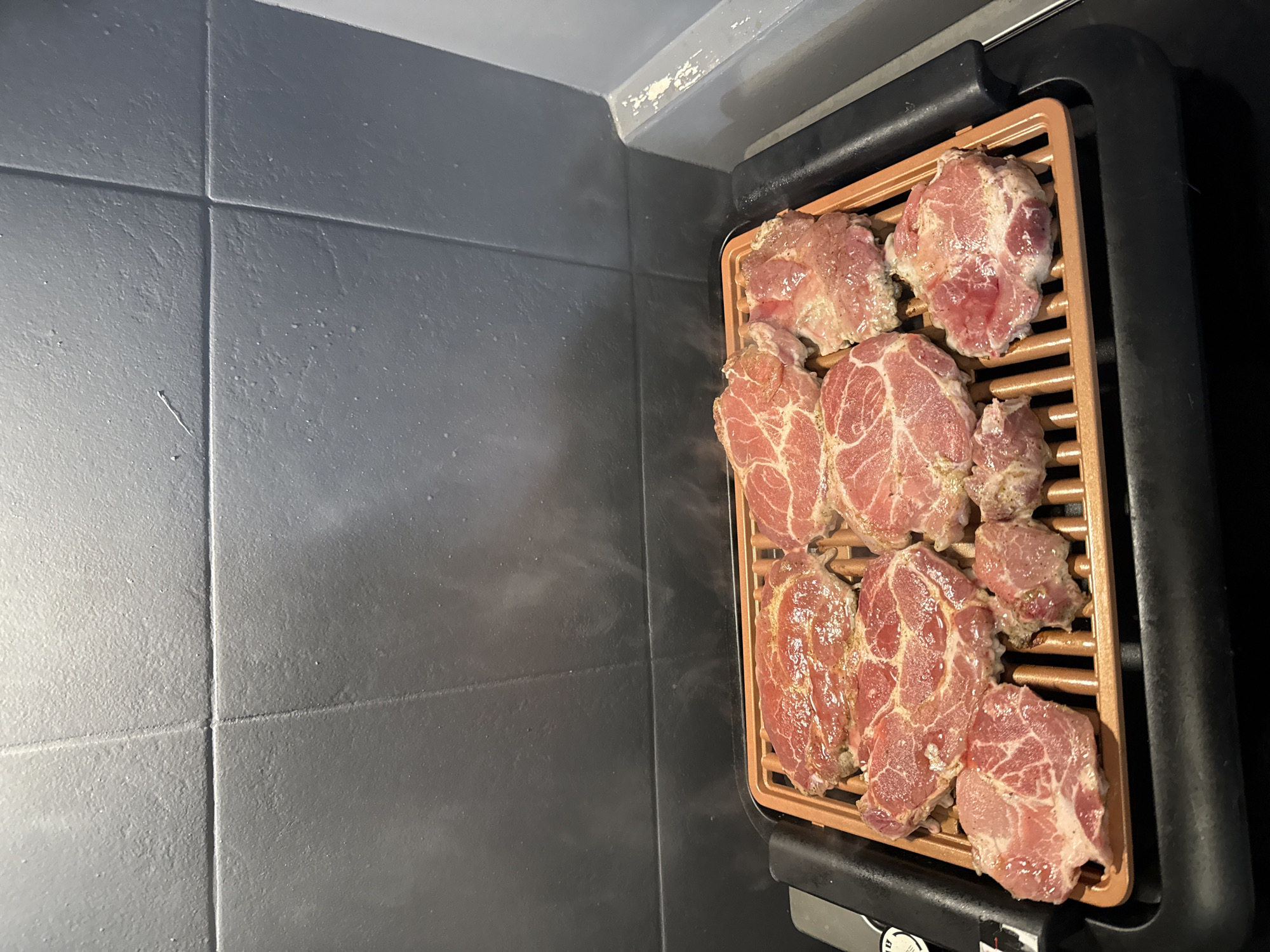 An overhead shot captures several raw, seasoned cuts of meat, likely pork neck steaks, resting on the copper-colored grates of an electric grill. The meat pieces are arranged for cooking, showing visible specks of seasoning. The grill has a black base and handles, with a control knob area visible at the bottom right, featuring partially legible white text, possibly including "OFF" and other settings. The background features a tiled wall composed of large, rectangular grey tiles. Above the tiled section, a strip of lighter grey or white surface is visible, showing some peeling paint or wear. To the right, a darker grey or black surface is present, possibly a countertop. The image depicts the pre-cooking stage in an indoor setting, situated in Stara Zagora, Bulgaria.
