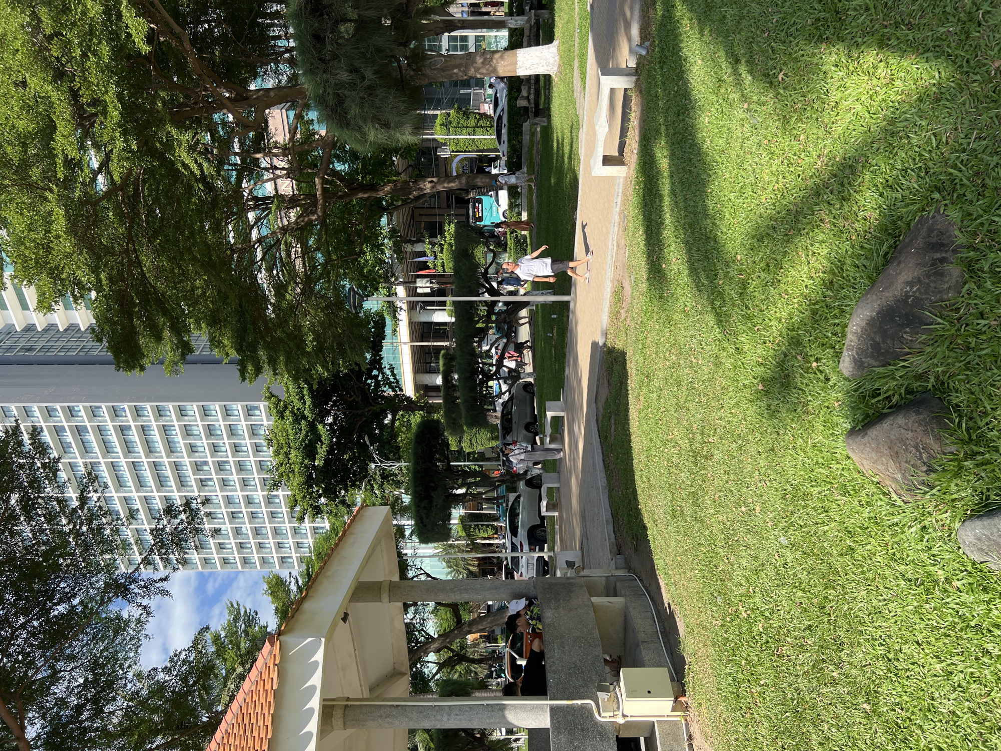 In Nha Trang, Vietnam, a sunny park scene unfolds. A young boy walks along a paved path, his shadow cast by the sun. To his right, a modern building with numerous windows rises high into the blue sky. Trees with lush green foliage are scattered throughout the park, providing shade. In the foreground, a patch of vibrant green grass is dotted with large rocks. To the left, a covered pavilion with a terracotta tiled roof offers a shaded seating area where several people are gathered. Cars are parked along the road in the background, and a turquoise bus is visible. The overall impression is one of a tranquil urban green space on a bright day.