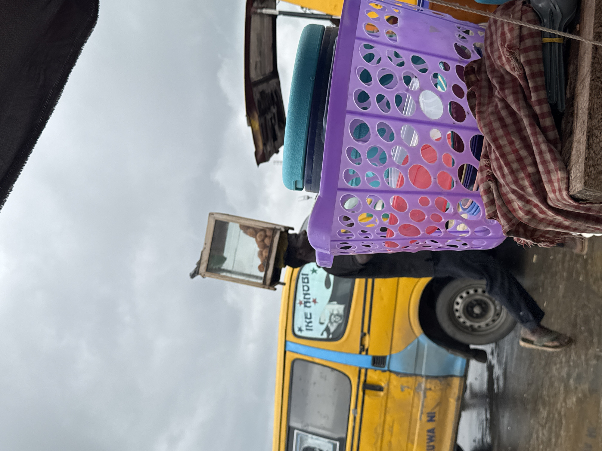A person is walking in front of a yellow and blue bus with the words "IKE ANOBI" written on it. The person is carrying a glass box filled with coconuts on their head. In the foreground, there is a purple basket filled with various items, including colorful plastic containers and some fabric. The ground is wet, suggesting it has recently rained or is still raining. The sky is overcast. The scene appears to be a street in Badagry, Nigeria, likely a market area given the presence of vendors and goods.