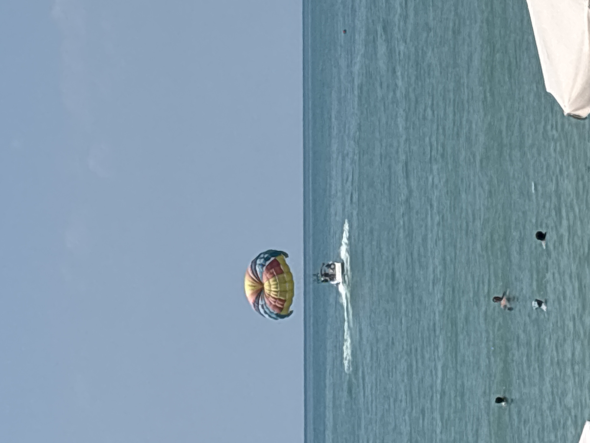 The image captures a sunny day at a beach in Sochi, Russia. The turquoise water stretches towards the horizon, meeting a clear blue sky. In the middle ground, a motorboat is actively pulling a colorful parachute carrying two individuals. The parachute is a vibrant mix of yellow, red, blue, and green, with its canopy inflated. In the foreground, several people are visible swimming in the ocean, their bodies partially submerged in the water. A white beach umbrella is partially visible in the bottom right corner, suggesting a recreational area. The overall scene depicts a typical summer day with water sports and beach activities.