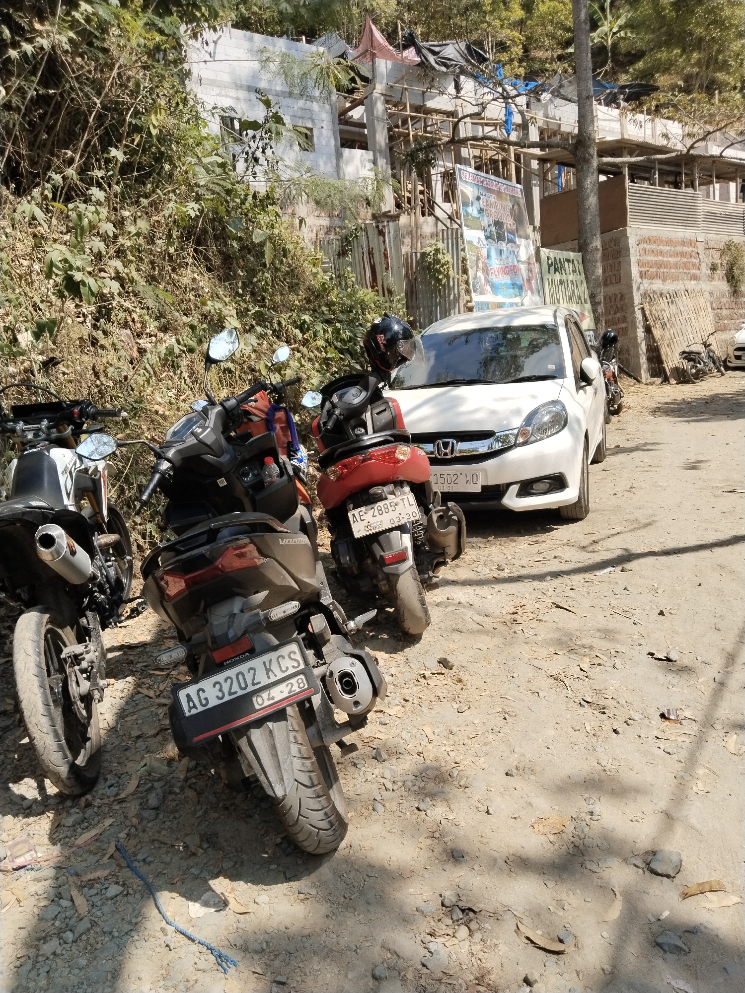 In the city of Prigi, Indonesia, a dirt road is lined with several parked motorcycles and a white car. The motorcycles, a mix of scooters and a dirt bike, are positioned in a row. A white car is parked behind them. The background reveals a building under construction, with scaffolding and building materials visible. Vegetation, including trees and bushes, surrounds the area, and a sign with text in Indonesian is posted on the building. The ground is covered in dirt and small stones, and sunlight casts shadows from the objects and trees.