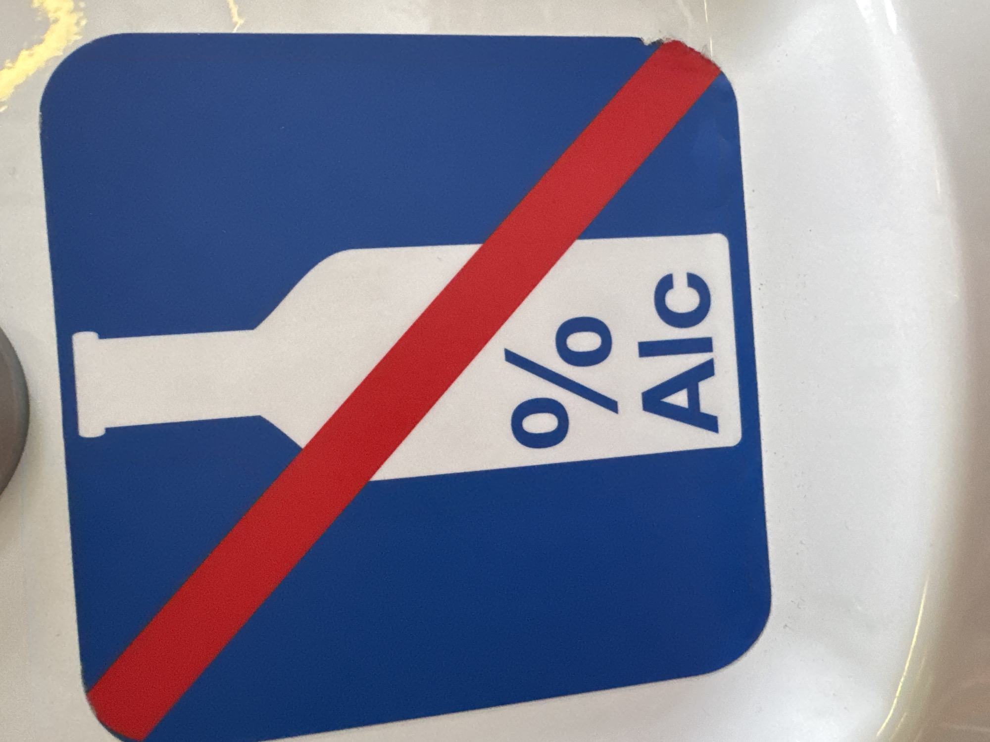 The image shows a close-up, slightly angled, vertical shot of a sign with a blue background and a white silhouette of a bottle with a red diagonal stripe across it. The words "%" and "Alc" are printed in white text on the bottle silhouette. The sign appears to be affixed to a white, curved surface, possibly a wall or a counter. The lighting suggests an indoor environment. The sign is a prohibition symbol indicating no alcohol.