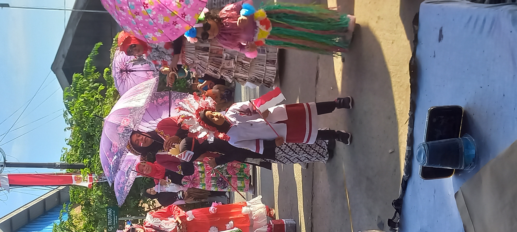 A group of people, including children and adults, are participating in an outdoor parade or celebration. The event appears to be a cultural or nationalistic display, as some individuals are wearing traditional Indonesian attire and carrying the Indonesian flag. The central focus is on a young child dressed in a white and red uniform, holding a small Indonesian flag. Other participants are seen holding decorative umbrellas and wearing colorful outfits. In the background, there are buildings and lush green trees. The overall scene suggests a festive atmosphere, possibly a local community event in Samarinda, Indonesia, given the presence of Indonesian flags and attire.