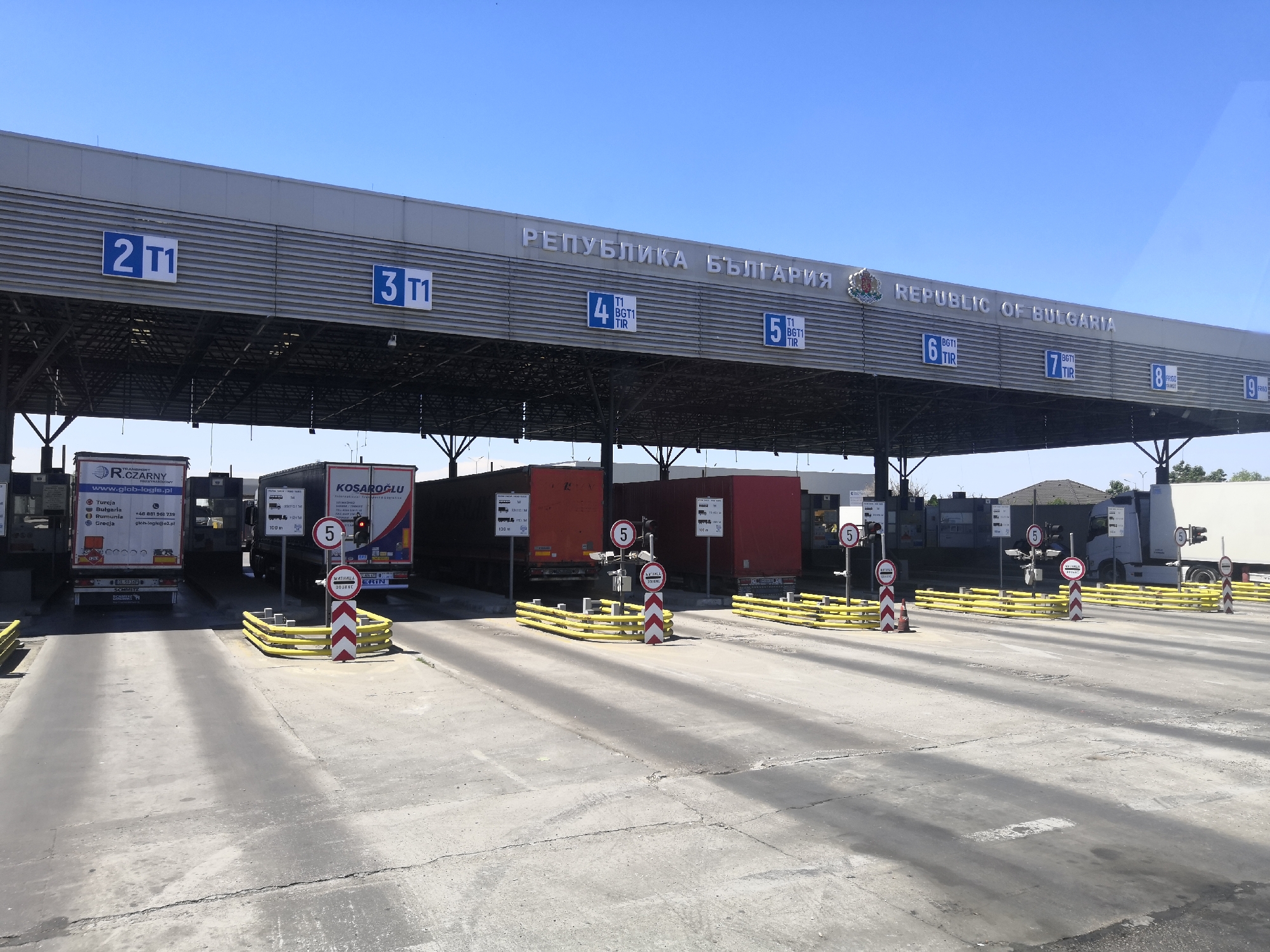 The image shows the Kapitan Andreevo border crossing in Bulgaria. The scene depicts a busy border checkpoint with multiple lanes for trucks. In the foreground, the concrete road surface is cracked and worn. Yellow metal barriers with red and white striped posts are present, directing traffic. Several trucks are visible, some stationary and some in motion, queuing at the different lanes. Above the lanes, a large structure with the text "РEПУБЛИKA БЪЛГAPИЯ" and "REPUBLIC OF BULGARIA" is visible, indicating the border crossing. Blue signs with numbers like "2 T1", "3 T1", "4 BGT1 TIR", "5 BGT1 TIR", "6 BGT1 TIR", "7 BGT1 TIR", and "8 BGT1 TIR" are mounted on the structure, denoting different lanes or checkpoints. One truck in the foreground has the branding "R. CZARNY" and a website address. Another truck further back has "KOSAROGLU" branding. The sky is clear and blue. The overall environment suggests a functional border crossing operation under daylight.