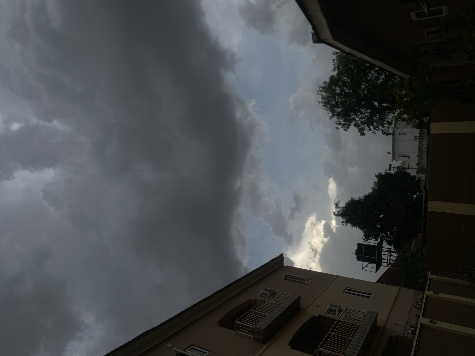 The image depicts an overcast sky above buildings in Barden Yana, Nigeria. The sky is dominated by dark, ominous storm clouds on the left, contrasting with lighter, brighter clouds on the right. Buildings are visible along the bottom and right edges of the frame. One building is beige with balconies and visible windows. Another building is taller and constructed from red brick. Trees are visible between the two buildings.