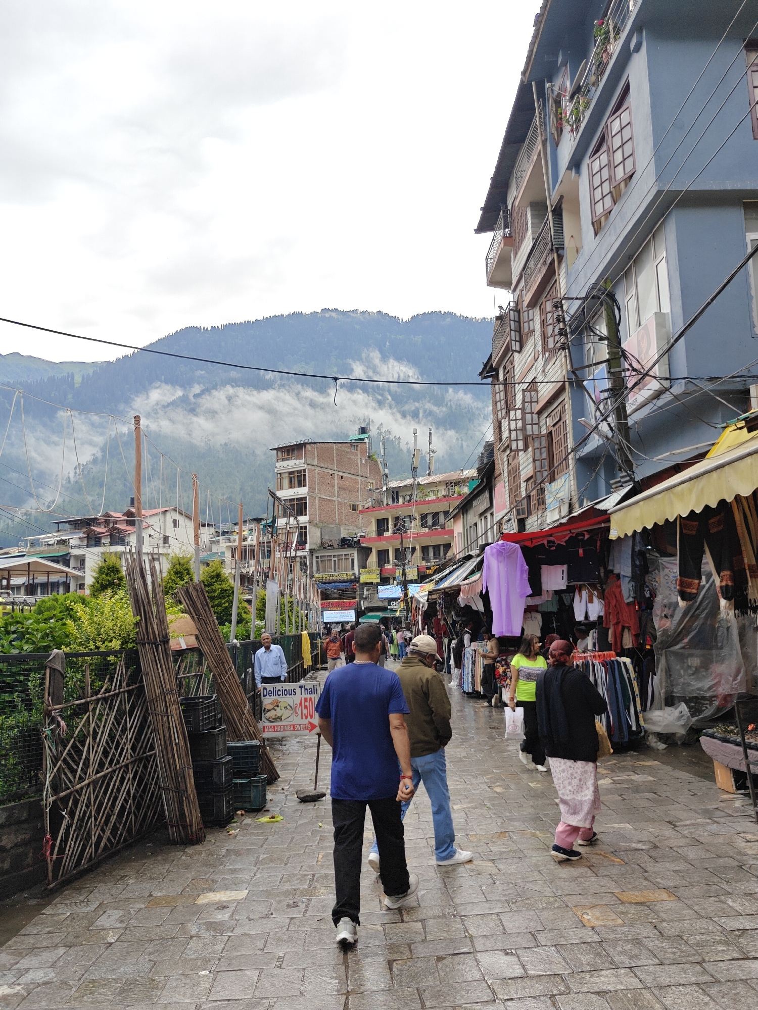 The image depicts a bustling street scene in Manali, India, likely on a cloudy day. In the foreground, several people are walking on a wet, paved street lined with shops and stalls. The architecture is a mix of traditional and modern buildings, with multiple stories and balconies visible. The background features a lush green mountain landscape partially obscured by mist, characteristic of Manali's scenic beauty.
Specifically, we see:
* **People:** A group of individuals, including men and women, are walking along the street. Some are casually dressed, while others appear to be shopkeepers or vendors. One man in a blue shirt is prominent in the foreground, walking away from the camera. A woman in a light pink patterned skirt and black top is also visible.
* **Shops and Stalls:** Numerous shops and temporary stalls are set up along the street, displaying a variety of goods, primarily clothing. Brightly colored fabrics and garments hang from racks, and some stalls have awnings for shade. A sign advertising "Delicious Thali" for ₹150 is visible.
* **Environment:** The street is paved with stone tiles that appear wet, suggesting recent rain or dampness. Wooden structures and bamboo poles are present, possibly part of the market setup or construction. The air seems cool and moist, with mist clinging to the mountains in the distance.
* **Objects:** Crate-like structures are stacked near a bamboo fence. Several electrical wires are strung across the scene, typical of urban environments in India.
The overall impression is one of a vibrant and active market area in a mountainous region.