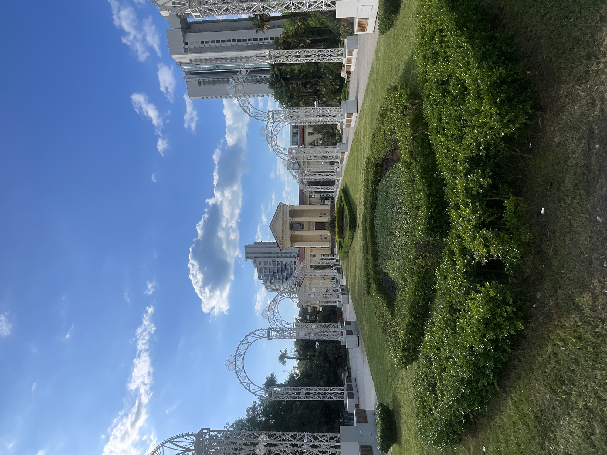 The image depicts a park in Sochi, Russia. A pathway is framed by a series of ornate white arches. The pathway leads toward a building with a tan facade. Green lawns and meticulously trimmed bushes flank the pathway. Tall buildings rise in the background. The sky is blue with scattered white clouds. The image appears to be taken during daylight hours. No people are clearly visible.