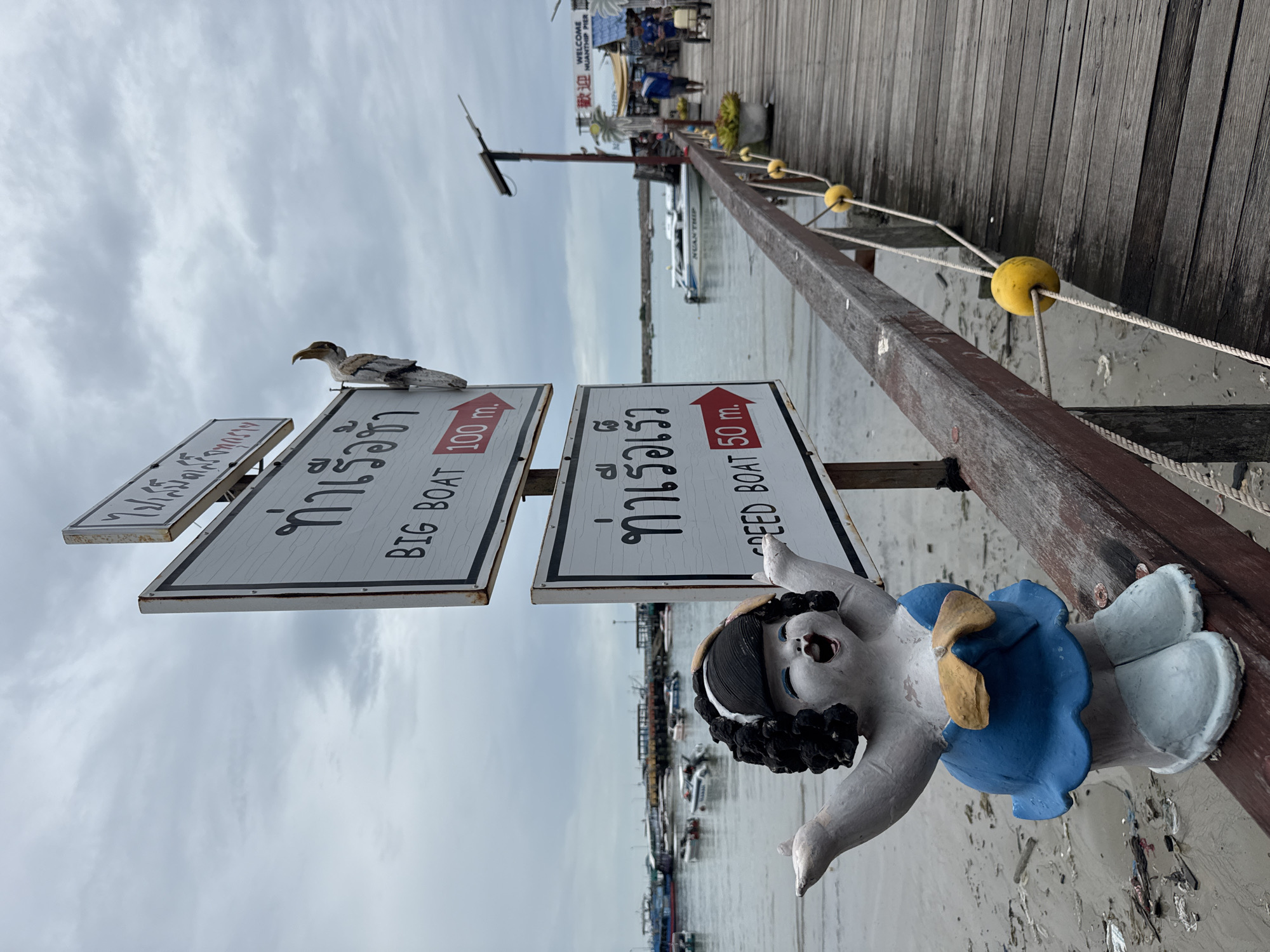 A statue of a joyful young girl in a blue dress stands on a wooden pier in Ban Phe, Thailand, with her arms outstretched. Behind her, a signpost directs visitors to "BIG BOAT" 100 meters to the right and "SPEED BOAT" 50 meters to the right. The pier extends into the water, where various boats are docked. The sky is overcast, and the water is calm. In the background, a sign reads "WELCOME NUANTHIP PIER," indicating the location. The scene is a typical harbor setting, bustling with activity and maritime elements.
