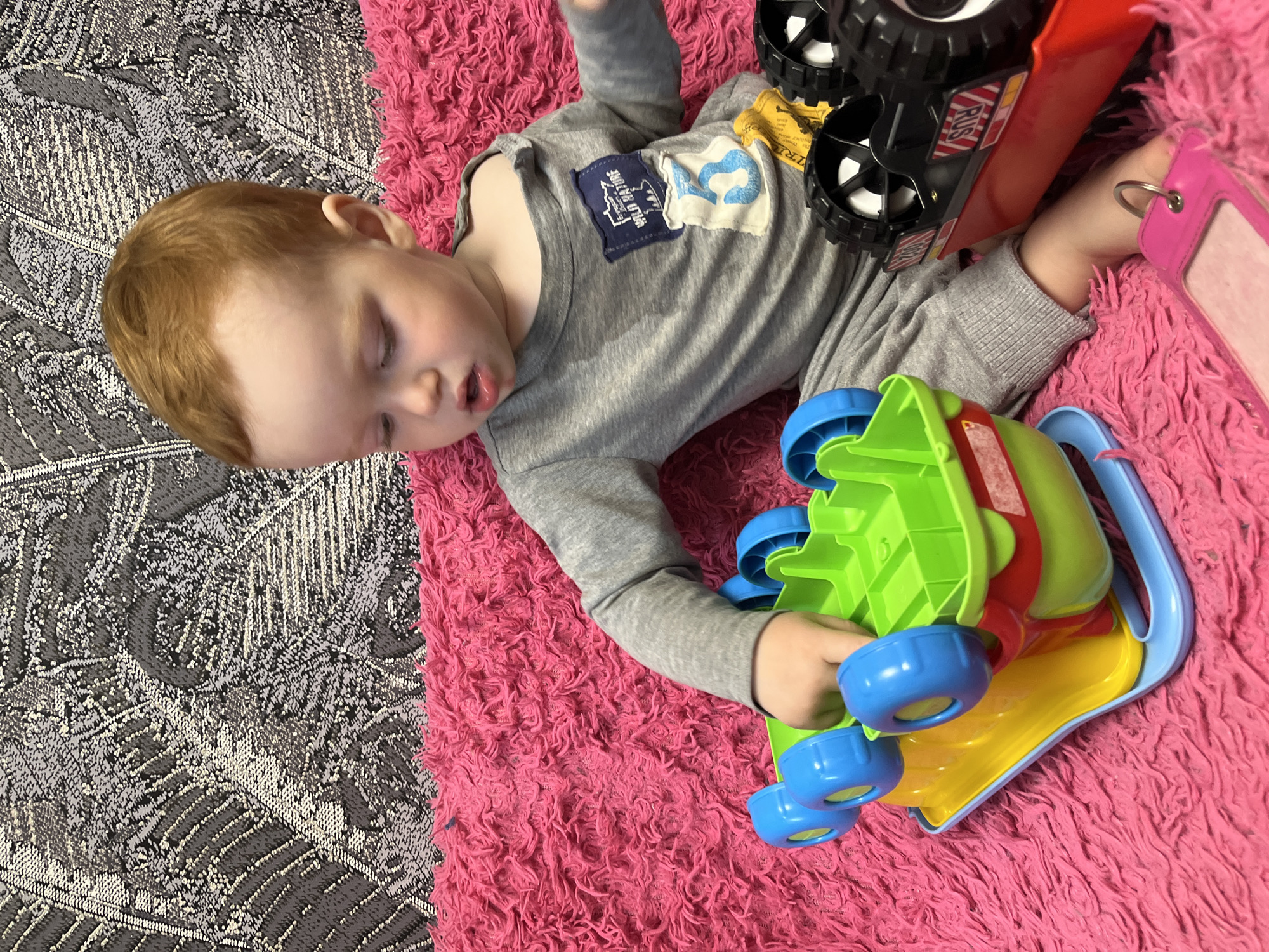 In a domestic setting, a young toddler with red hair is seated on a bright pink, plush surface, possibly a couch or rug. The child is wearing grey pajamas and appears engaged with two colorful toys. To the left, a green and yellow toy vehicle with blue wheels is positioned in front of the child, with the child's hand touching it. To the right, a black and red toy truck with large wheels is also present. The background features a patterned fabric, with a leaf-like design in shades of grey and white. The image is well-lit, with a naturalistic feel. No information is available regarding the specific location within the city of Chaikovsky, Russia.