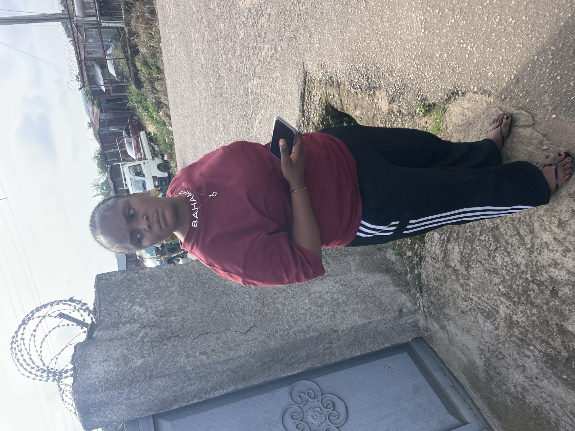 A medium shot captures a Black woman standing on a paved area next to a rough-textured concrete wall. She is facing right, looking away from the camera. The woman wears a maroon t-shirt with white text that appears to spell "BAHAMAS" and black track pants with white stripes down the sides. She holds a dark-colored smartphone in her right hand. The background shows a street with several vehicles, including a white truck and a red car, and a cluster of corrugated metal structures. The sky is overcast. The concrete wall on the left has barbed wire coiled along its top. The ground consists of asphalt and some dirt with sparse vegetation.