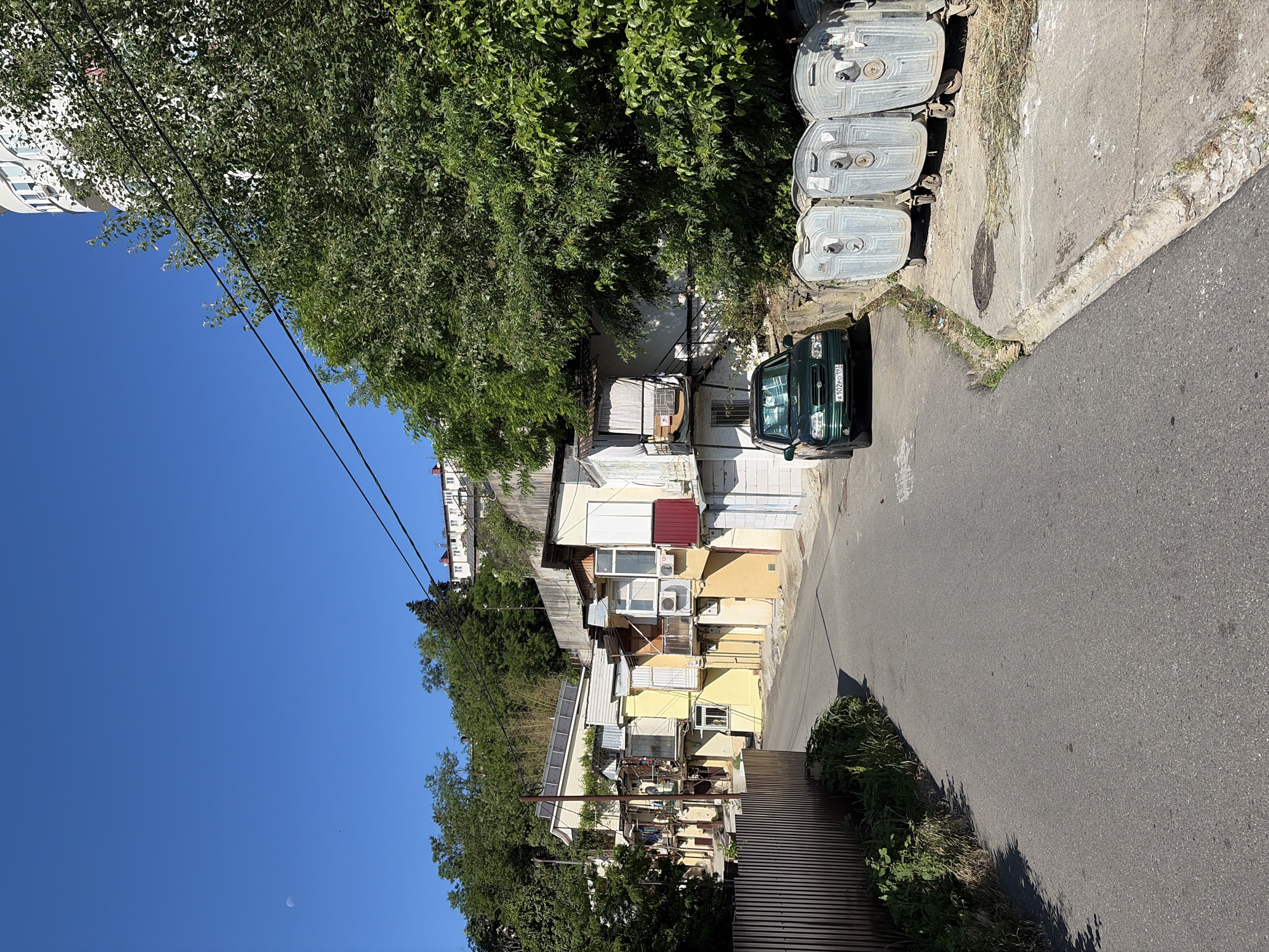 This detailed description pertains to an image taken in Sochi, Krasnodar Krai, Russia.
The image presents a residential street winding through a steep, verdant hillside under a clear, bright blue sky with a visible crescent moon in the upper left. The time of day appears to be morning or late afternoon, indicated by the high sun and distinct shadows.
In the foreground and midground, a paved asphalt road curves gently to the left. On the left side of the road, a dark brown corrugated metal fence borders a lush bank of green foliage. On the right side of the road, a concrete curb separates the asphalt from a concrete sidewalk. Three large, grey, rectangular garbage bins with wheels and distinctive patterned sides are lined up along this sidewalk. Next to the bins and facing the viewer, a dark green SUV (likely a Suzuki Grand Vitara) is parked. Its Russian license plate reads "A 102 PO 193," confirming its registration in the Krasnodar Krai region. A circular manhole cover is visible on the sidewalk between the car and the bins.
Along the left side of the street, a series of multi-story residential buildings are constructed directly into the steep slope. These houses exhibit varied architectural styles and colors, including shades of yellow, cream, and white, suggesting a mix of original construction and subsequent additions or renovations. Several feature small balconies, windows with exterior blinds, and exposed air conditioning units. One prominent house in the mid-left has multiple solar panels installed on its roof.
In the background, atop the highest point of the hill, several white buildings with red roofs are discernible. Higher on the right side of the image, a large, modern-looking white apartment building with curved exterior walls and multiple windows is partially visible, obscured by dense green tree canopy and two prominent black power lines that traverse horizontally across the upper right quadrant. The overall scene depicts a densely populated, hilly urban environment characteristic of the Black Sea coastal city. No people or animals are visible in the scene.