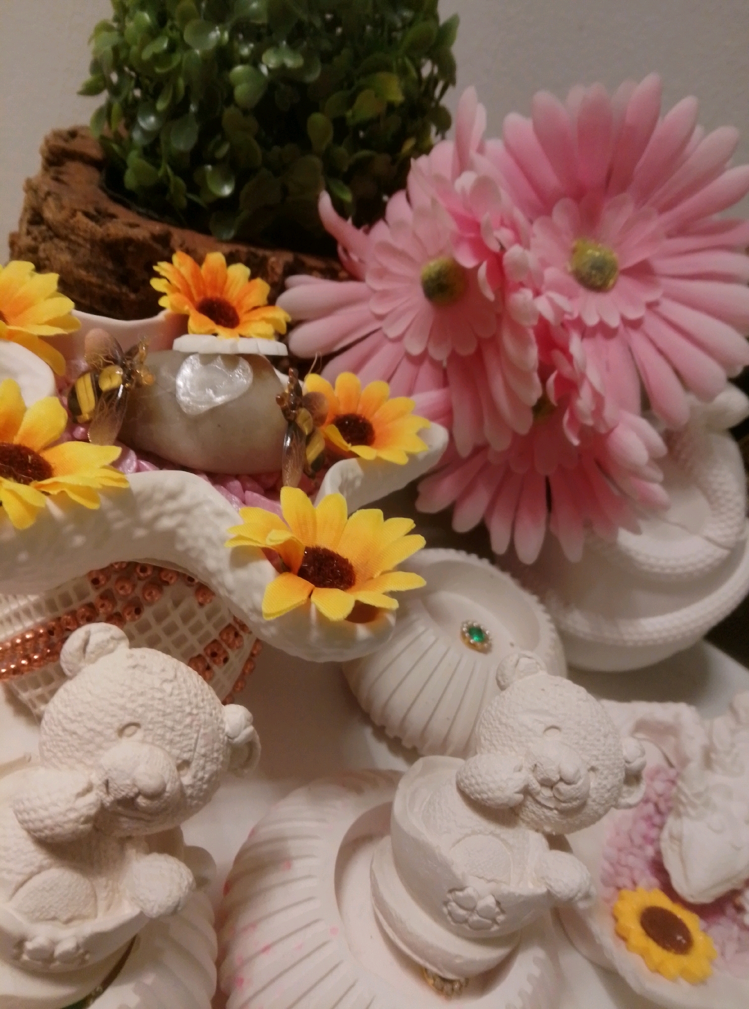 The image shows a close-up of a decorative arrangement with a whimsical theme. In the foreground, there are two white, textured bear figurines. The bear on the left is sitting with its arms up to its face, and the bear on the right is in a more recumbent position, also appearing to hold its arms. Surrounding the bears and other decorative elements are various artificial flowers, primarily sunflowers with yellow petals and dark centers, and large pink gerbera daisies. There are also small, decorative bee figurines scattered throughout the arrangement, as well as some white shell-like decorative pieces. In the background, a cluster of artificial green foliage rises from behind a piece of textured brown wood. The overall impression is of a playful and decorative display, possibly a cake topper or a table centerpiece. The lighting is soft, and the focus is on the intricate details of the various components. There is no discernible location in the city of Wedel, Germany depicted in this image.