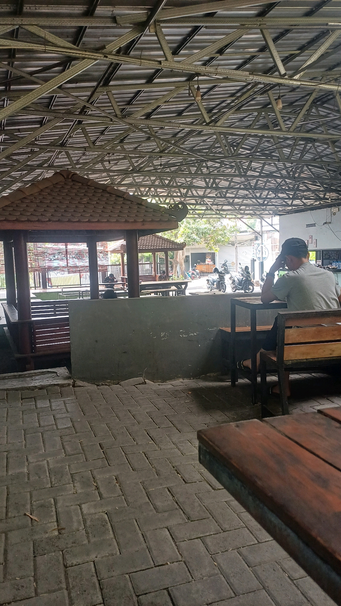 The image depicts an outdoor seating area with a roof structure supported by metal beams. A person wearing a gray t-shirt and a black baseball cap is seated at a wooden table and chair, facing away from the camera and resting their head on their hand. The ground is paved with interlocking brick pavers. In the background, a traditional gazebo-like structure with a tiled roof is visible, along with other seating areas, trees, and motorcycles parked on a street. The environment appears to be a commercial establishment, possibly a cafe or restaurant, located in Tlogopatut, Indonesia. The lighting suggests it is daytime, with shadows indicating the sun's position.