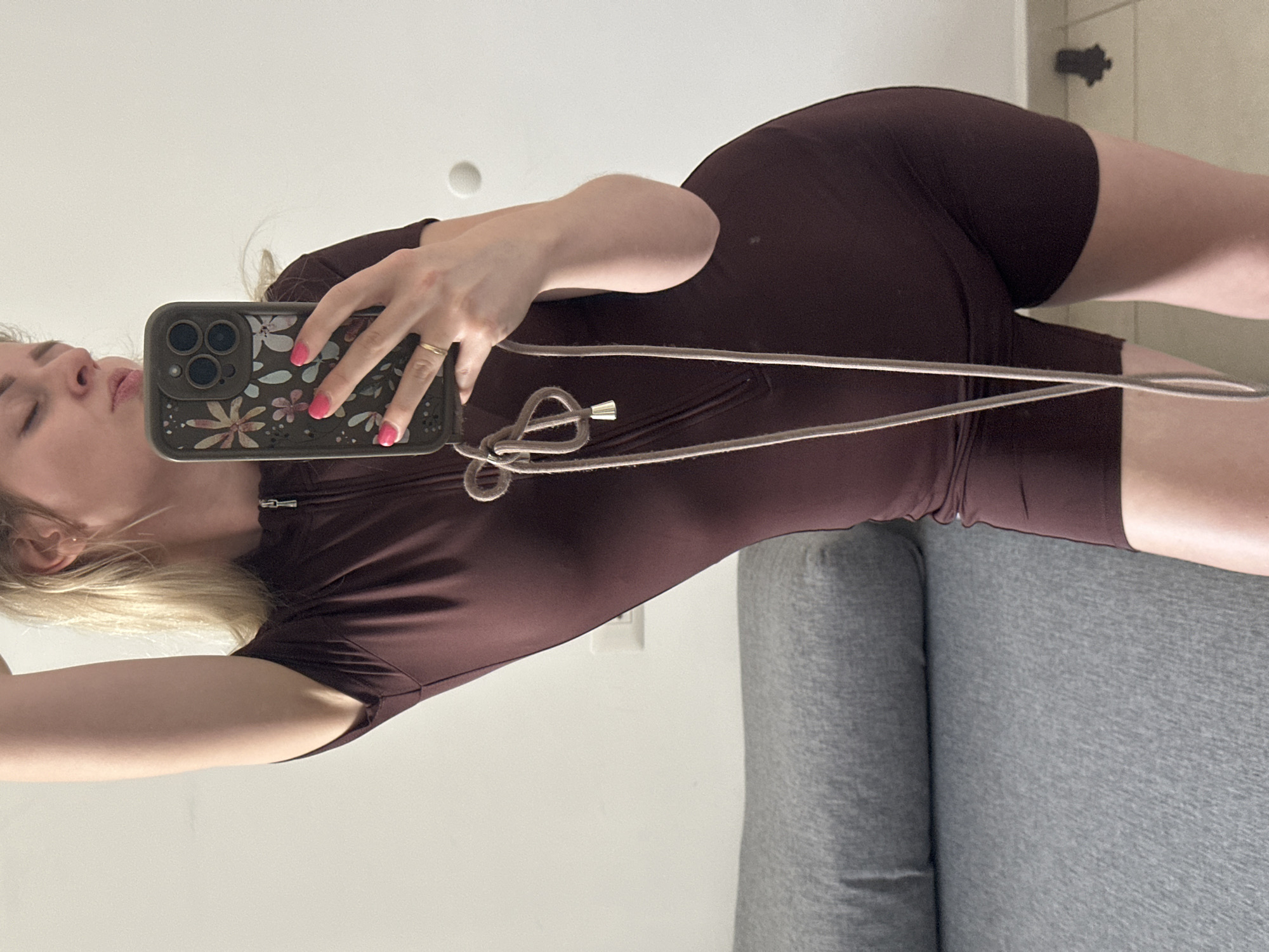 An indoor mirror selfie captures a young woman posing in a room. The image is rotated 90 degrees clockwise, showing the woman horizontally. She has blonde hair and light skin, and her lips are slightly pursed. Her right hand holds an iPhone, which is equipped with a dark green or grey floral-patterned case featuring prominent camera lenses and an attached lanyard-style strap. Her fingernails are painted pink, and she wears a simple gold-colored ring on her left ring finger.
The woman is wearing a dark brown, form-fitting, short-sleeved and short-legged garment, possibly a jumpsuit, playsuit, or bodysuit. This outfit has a zipper detail on the front and a decorative drawstring tied at the chest or waist area. Her left arm is slightly raised. In the background, a plain white wall is visible, along with a circular light fixture or object. A portion of a grey upholstered couch or sofa can be seen on the right side and bottom of the frame, providing a soft contrast to the stark white wall. No visible text is present in the image.