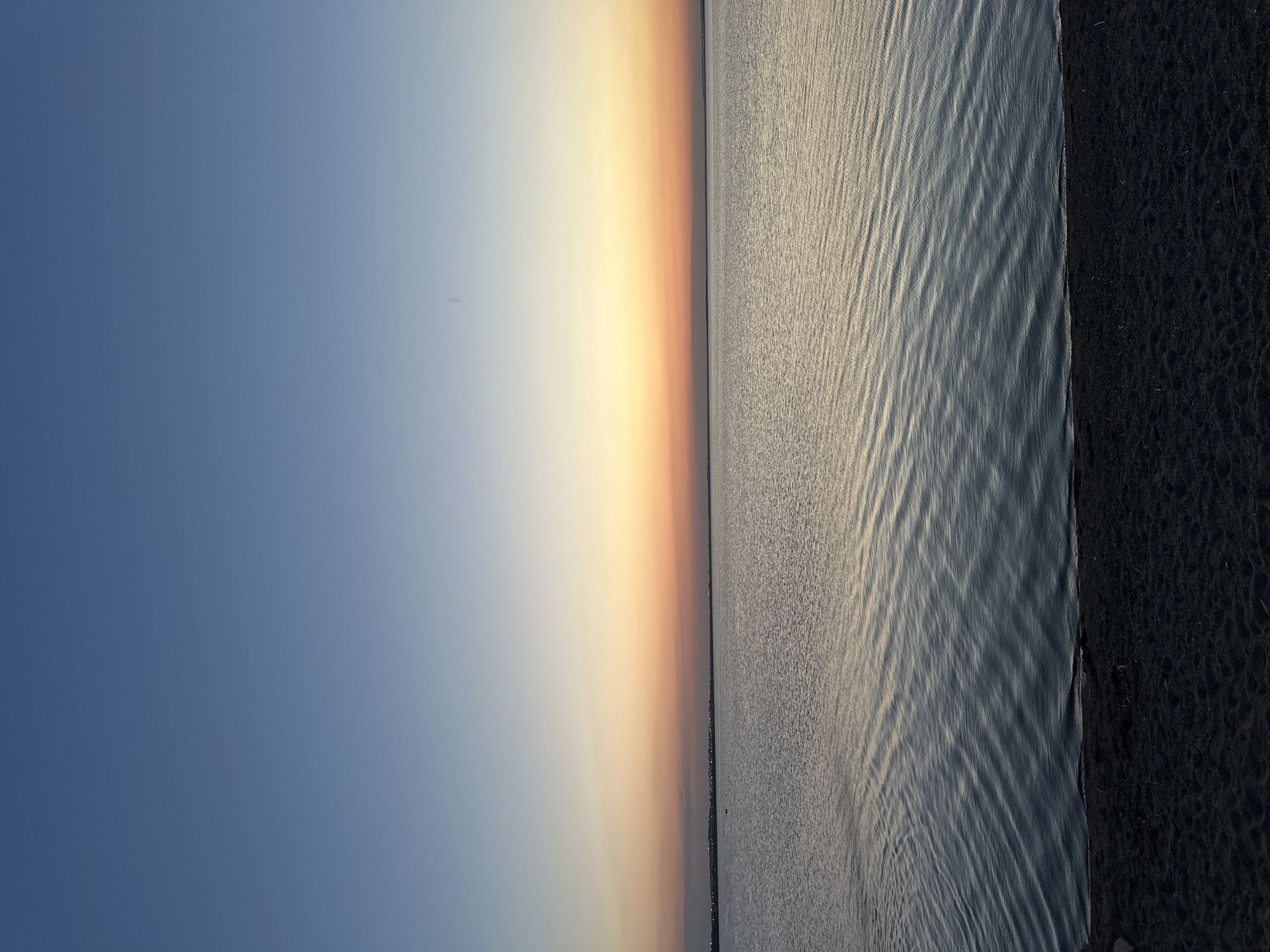 The image depicts a serene body of water, likely a lake, bordering a dark, sandy shore. The sky above transitions from a deep blue at the top to a warm gradient of orange and yellow near the horizon, suggesting either sunrise or sunset. A faint line of lights is visible on the distant shore, indicating the presence of a settlement or city. The water is calm, with gentle ripples reflecting the colors of the sky. The overall scene is one of natural tranquility and the beauty of twilight. This could be a location along the shores of Lake Borovoe, near the city of Kokshetau, Kazakhstan, known for its picturesque natural landscapes and the opportunity to witness such atmospheric phenomena.