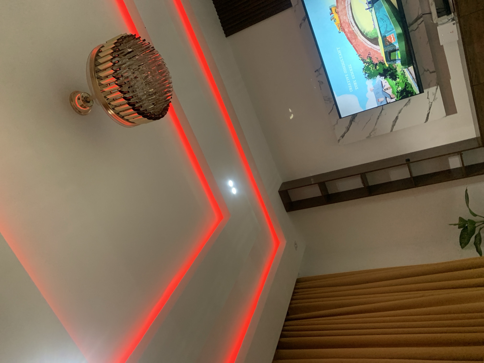 The interior shows a wall-mounted light fixture with a golden finish on the left side of the frame. To the right are two vertical lines of red lights. A television set is mounted on the wall, slightly off-center towards the upper right. The television displays an animated scene with text visible. Below the television is a wooden shelf with several compartments. Partially visible in the bottom right corner are brown window blinds and the top of a green plant.
There is nothing in this image to indicate it is in Jos, Nigeria.
