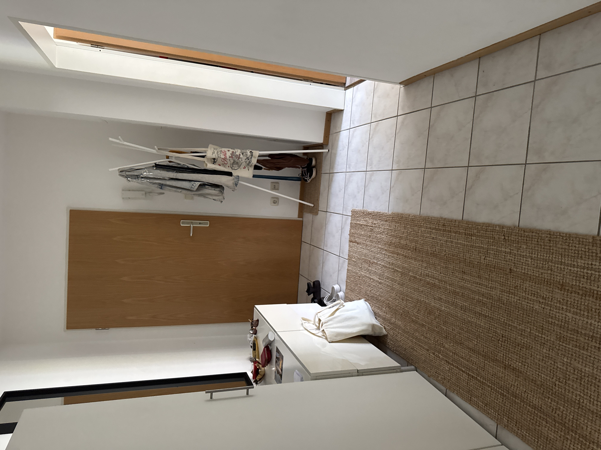 The image depicts an indoor hallway in an apartment located in Höhenkirchen-Siegertsbrunn, Germany. On the left, a white cabinet with a drawer is partially visible, topped with various items including a red bowl, some papers, and other miscellaneous objects. A white tote bag rests against the cabinet, and a woven rug runs along the floor in front of it. Further into the hallway, a closed wooden door is present. To the right of the door, a white coat rack stands with several items of clothing and a patterned tote bag hanging from it. Two pairs of shoes are visible on the floor near the coat rack, one being black sneakers and the other appearing to be lighter colored casual shoes. The floor is tiled with a light-colored, square pattern. The walls are painted white, and a portion of a doorway leading to another room is visible on the far right of the image, with another wooden door partially ajar. The lighting suggests an indoor environment with natural light coming from the right.