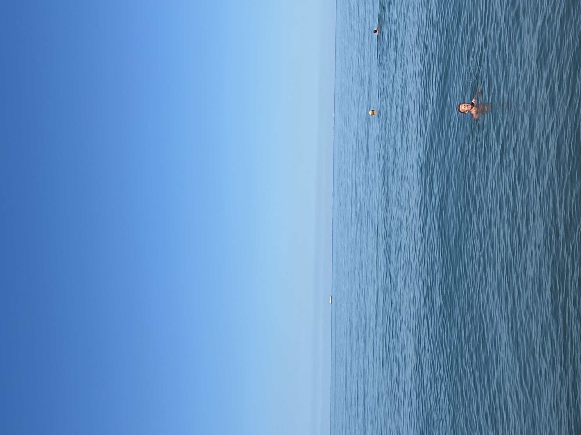 A man is swimming in the Black Sea off the coast of Sochi, Russia. The sky is clear blue and the water is calm with small ripples. In the distance, there are two small boats on the horizon and a yellow buoy. Another person can be seen swimming further away from the main subject. The man in the foreground is looking at the camera.