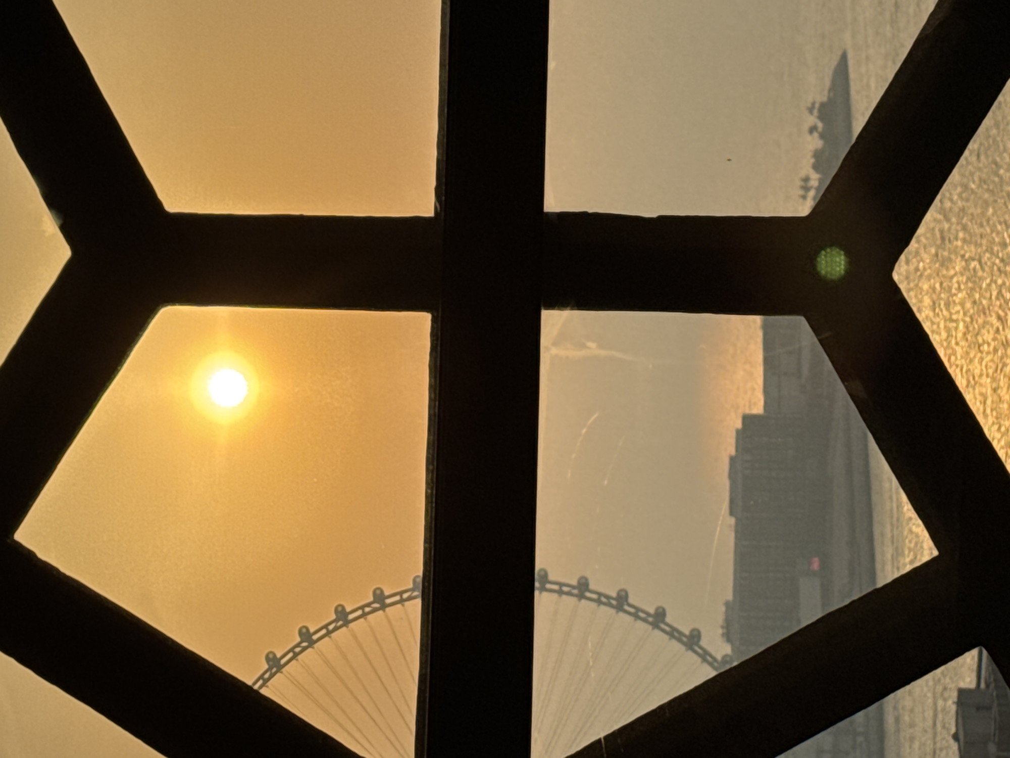 The image depicts a sunset view through a window frame in Dubai, United Arab Emirates. The sun is a bright orb in the upper portion of the frame, casting a golden glow. A Ferris wheel, likely Ain Dubai, is visible on the left, with its spokes extending towards the sun. In the middle ground, a large building complex with a red light on its facade is situated on the waterfront. The sea stretches to the horizon, reflecting the sunset's colors. A small island with palm trees is visible on the right. The foreground is dominated by a dark, geometric window frame, which divides the view into several sections. The scene is serene and bathed in the warm hues of dusk.