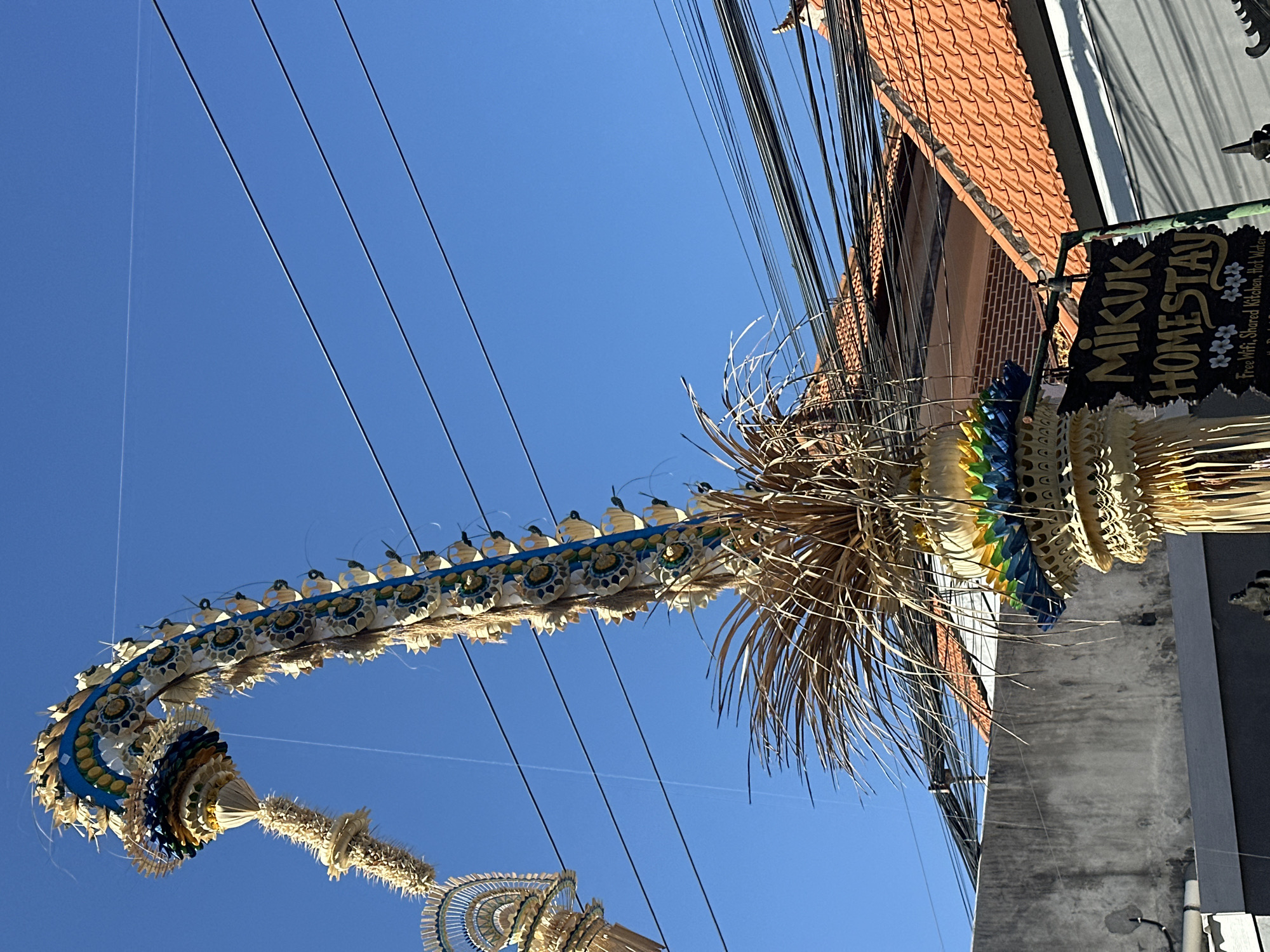 This image captures a detail of a tall, elaborate decorative structure, likely a Penjor, made of natural materials such as dried leaves and woven palm fronds, adorned with blue and yellow elements. The Penjor curves gracefully against a clear blue sky. Below, a tangle of black electrical wires stretches across the frame, crisscrossing in front of buildings with orange tile roofs. A sign for "MIKUK HOMESTAY" is visible, also made of natural materials, indicating the location is likely a street in North Kuta, Indonesia. The scene is bathed in sunlight, creating strong shadows and highlights.