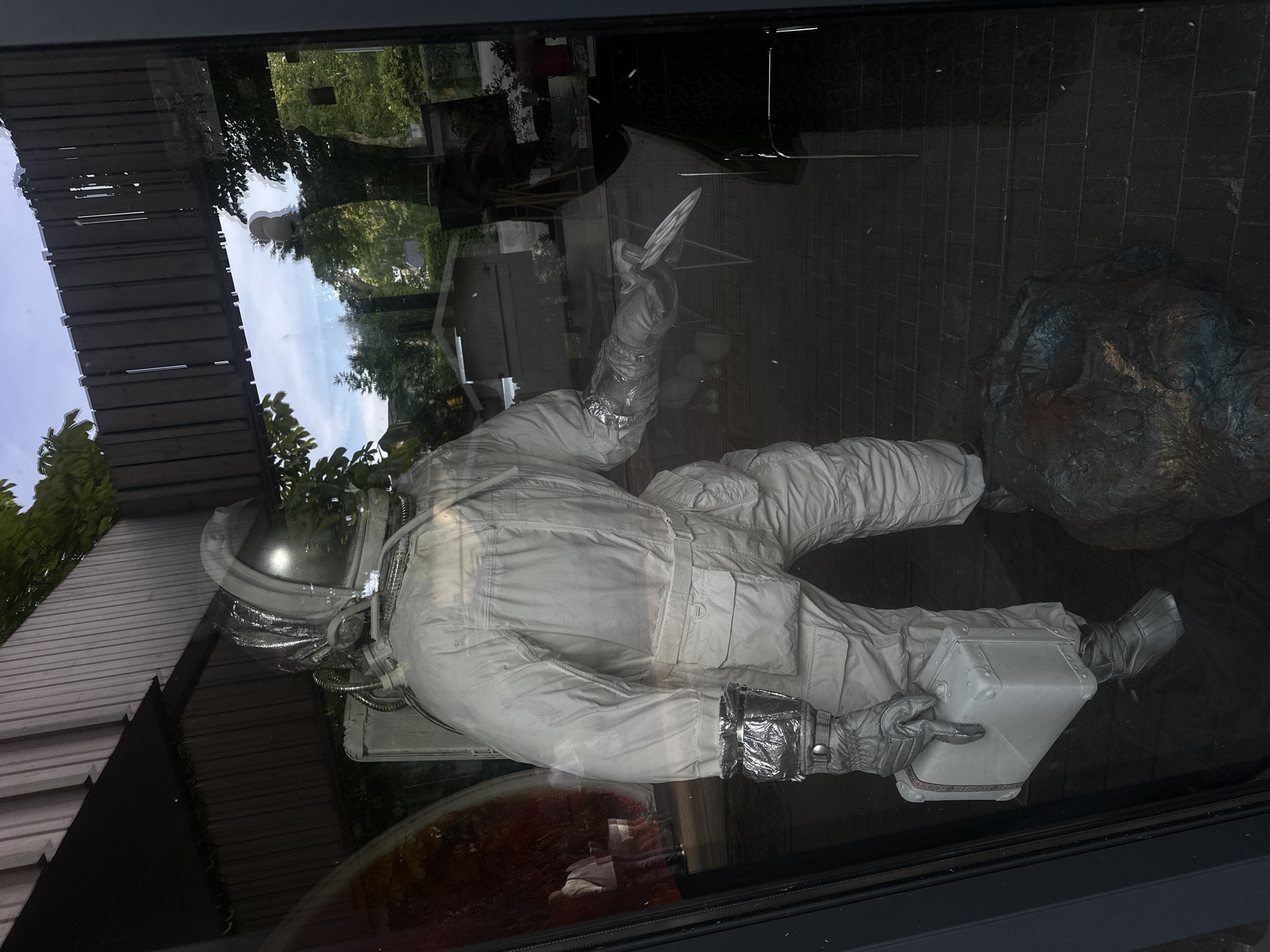 The image shows a mannequin dressed as an astronaut standing in a window display. The astronaut is wearing a white, bulky suit with reflective material on the helmet, gloves, and boots. The mannequin is holding a small, rectangular suitcase in its left hand and a sharp object, possibly a knife, in its right hand. In front of the mannequin, on the ground, is a dark, textured object that appears to be a sculpture or prop. The background visible through the window shows greenery, trees, and a bright sky, suggesting an outdoor environment. Reflections on the glass indicate the presence of buildings and a wooden walkway overhead. The overall scene is dimly lit, with light glinting off the astronaut's suit. This display is located in the city of Jūrmala, Latvia.