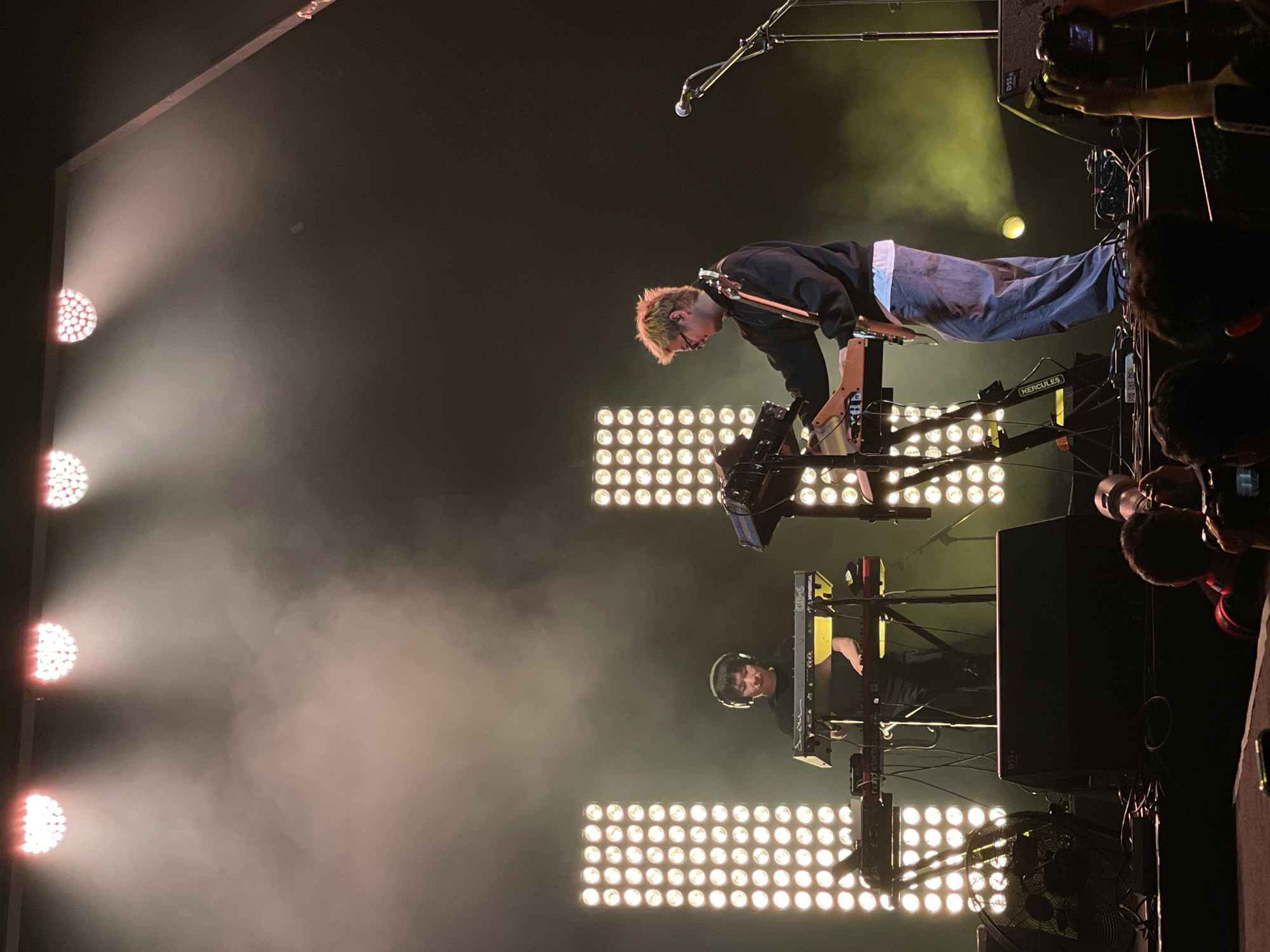 The image depicts a musical performance, likely a concert, with stage lighting creating a hazy atmosphere. In the foreground, a musician with blond hair is bent over, playing an instrument resembling a keyboard or synthesizer on a stand. The musician is wearing a dark jacket, and light-colored pants. Another musician is visible in the background, also playing a keyboard or synthesizer, and wearing headphones. The musicians are positioned in front of a large array of bright stage lights. Audience members are visible on the right side of the frame, holding up cell phones to record the performance. The scene is likely indoors, based on the controlled lighting and the lack of natural light. The location is Salembaran, Indonesia, but this information is not evident in the image alone.