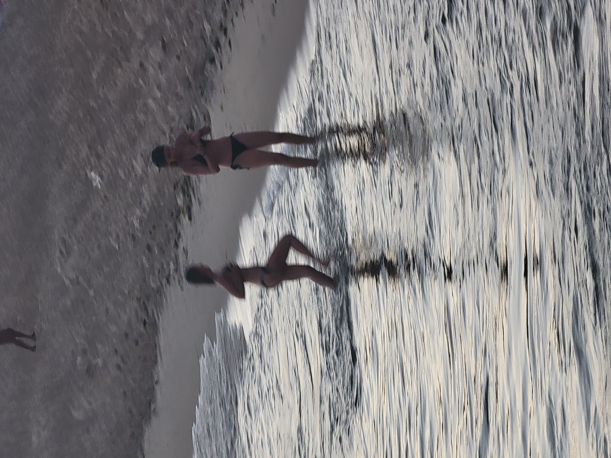Two women are partially submerged in shallow water at the beach in Quarteira, Portugal. The woman on the left, closer to the foreground, is facing away from the camera, with her legs visible in the water and her arms raised slightly. The woman on the right, further back and on the right side of the image, is facing the camera, wearing a dark bikini and a dark cap, and holding something in her hands. Both women are wearing dark bikinis. The water is rippling, reflecting the muted tones of the sky and sand. The beach sand is visible behind the women, sloping upwards. Another person's legs are visible standing on the higher sand bank in the upper left portion of the image. The overall scene is a candid moment captured at the water's edge.
