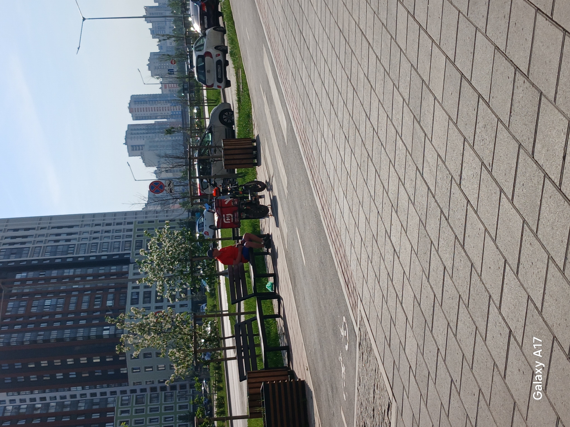 The image depicts an outdoor scene with a long stretch of paved walkway dominating the right side of the frame, marked with brick-like patterns. Adjacent to the walkway is a narrower paved path with white directional arrows and designated spaces for bike parking. On the left side of the path, a row of wooden benches are positioned, some with wooden bin enclosures beside them. A person wearing a red shirt and blue shorts is seated on one of the benches. A red delivery scooter is parked adjacent to the benches. Behind the benches is a strip of grass with small trees and a view of a tall apartment building with many windows. In the distance, other high-rise buildings can be seen under a clear blue sky. The location appears to be in the city of Yekaterinburg, Russia.
