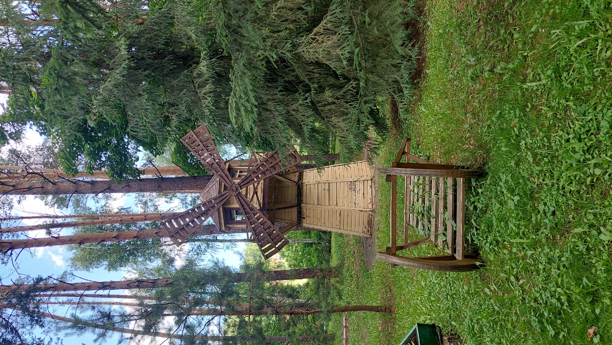 In Potashnaya Polyana, Russia, a rustic wooden windmill stands amidst a grove of tall pine trees and dense green foliage. The windmill, painted a faded yellow, features wooden blades and a small windowed upper section. In the foreground, a weathered wooden structure, possibly a decorative bridge or part of a cart, rests on the grassy ground. The scene is bathed in natural daylight under a partly cloudy sky.
