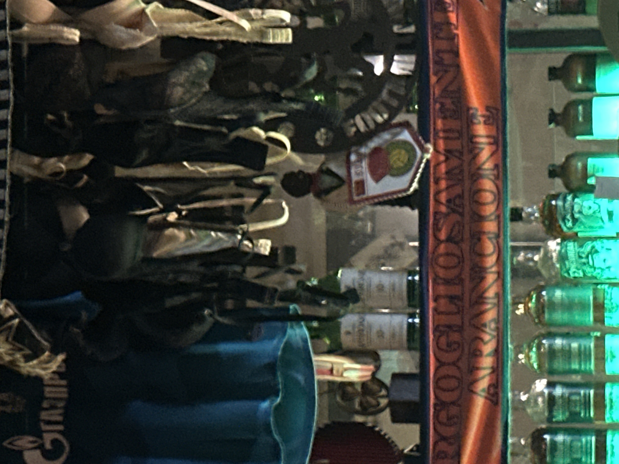 The image depicts a shelf stocked with various items, including bottles of alcohol and decorative objects. In the background, there are several hanging items that appear to be fabric or clothing. A banner with text in Italian, "ORGOGLIO ARANCIONE," is displayed across the middle of the shelf. On the left side of the image, a logo with Cyrillic letters "ГАЗПРОМ" is visible. The scene is dimly lit, with some bottles emitting a green glow from underneath. The context suggests this is a bar or a retail establishment. It is not possible to determine the specific location in the city of Minsk, Belarus from the provided image.
