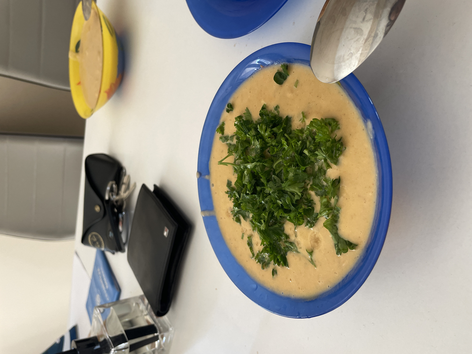 On a white surface, presumably a table, is a bowl of creamy soup garnished with fresh parsley. The soup is served in a blue bowl. A spoon rests partially within the bowl, likely used for consumption. To the left of the bowl, a black wallet sits beside a glasses case containing Ray-Ban sunglasses. Beneath these items is a clear container holding what appears to be writing utensils. A partially visible yellow bowl sits above the main bowl. The scene is lit with natural light. There is no indication of this image being taken in Plovdiv, Bulgaria.