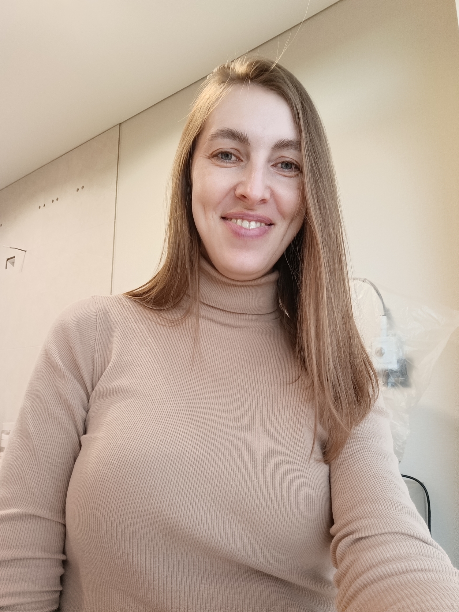A woman is centered in the frame, smiling directly at the camera. She has fair skin, blue eyes, and light brown hair parted to the side. She wears a tan turtleneck sweater. The background is a neutral color with a partially visible electrical outlet covered in clear plastic. The location is not discernible from the image. There is no indication the photo was taken in Perm, Russia.