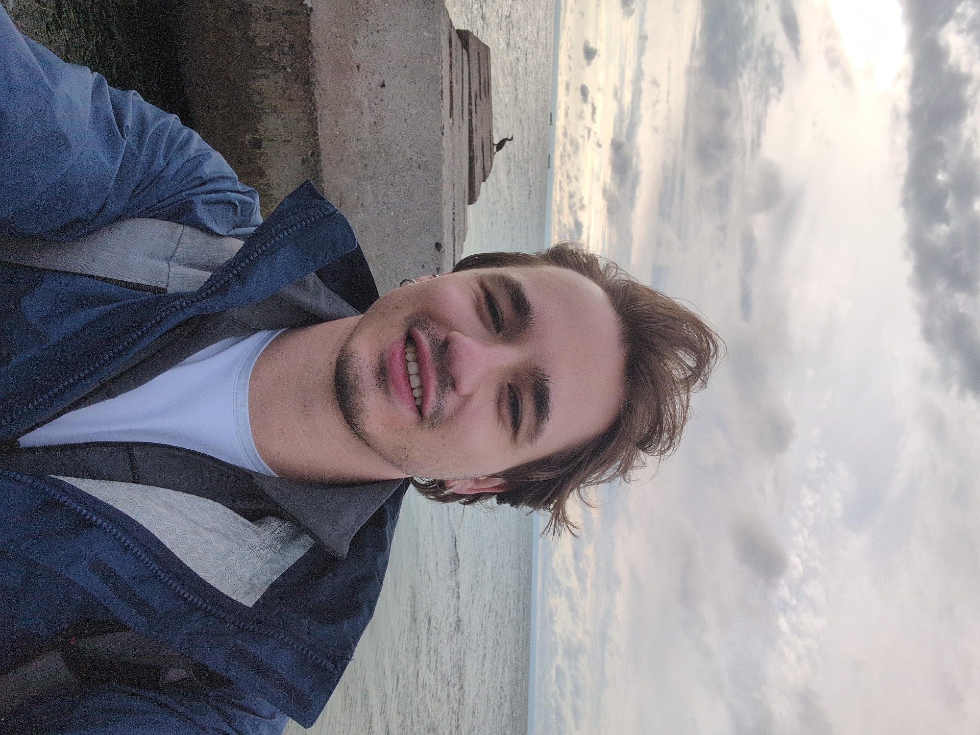 A young man with light brown hair, a light blue t-shirt, and a dark blue jacket takes a selfie by the sea. Behind him, the sea stretches to the horizon under a cloudy sky. To his left, a concrete pier extends into the water, with a bird standing on it. Several ships are visible in the distance on the water. The man is smiling slightly, with a small beard and a hoop earring in his left ear. The image appears to be taken outdoors, with the sea and sky as the background. The location is Odesa, Ukraine.