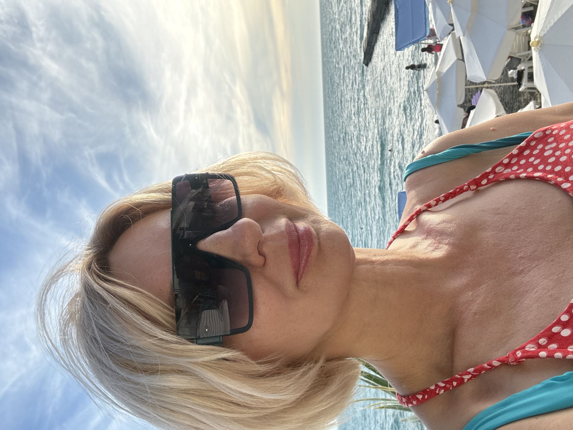 A woman with blonde hair and sunglasses takes a selfie on a beach. She is wearing a red and white polka dot bikini with a turquoise strap. The ocean is visible behind her, with some beach umbrellas and people in the distance. The sky is cloudy and bright, suggesting it is daytime. The image appears to have been taken in Sochi, Russia. The woman's expression is neutral, and the overall mood of the photo is relaxed and summery. There is no visible text in the image.