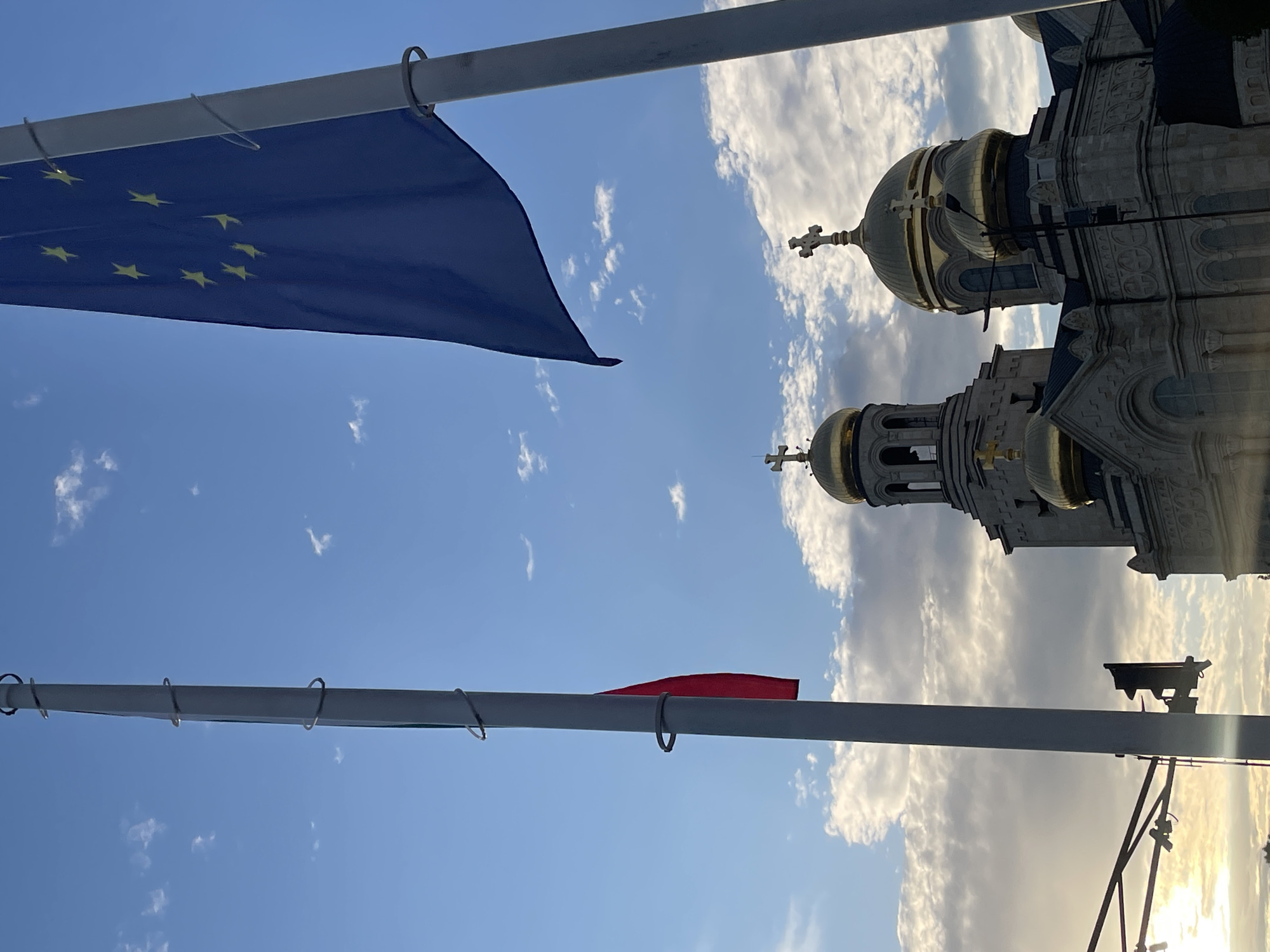 Two flagpoles stand vertically against a bright blue sky with scattered white clouds, indicating a daytime scene. The left flagpole displays a section of a national flag, revealing vertical red and a glimpse of green fabric, consistent with the lower portion of the Bulgarian national flag. The right flagpole displays the flag of the European Union, characterized by its dark blue field with twelve golden stars arranged in a circle.
Below and between the flagpoles, the upper section of the Dormition of the Theotokos Cathedral (Assumption Cathedral) in Varna, Bulgaria, is prominently featured. The cathedral exhibits multiple ornate golden domes, each surmounted by a cross. Two large domes are distinctly visible, along with a taller, bell tower-like structure to the left, also capped with a golden dome and cross. The cathedral's facade is constructed from light-colored stone, adorned with intricate architectural detailing. Due to the low-angle sun on the left, the building is partially silhouetted, with the golden domes reflecting the light.
The sky transitions from a lighter blue with wispy cirrus clouds at the top to a more textured and partially overcast sky near the horizon on the left, where a strong golden light from the low sun illuminates the clouds. A dark, partial silhouette of a street light or traffic signal pole is visible on the far left edge of the frame, positioned against the bright light source. No individuals are present in the image.