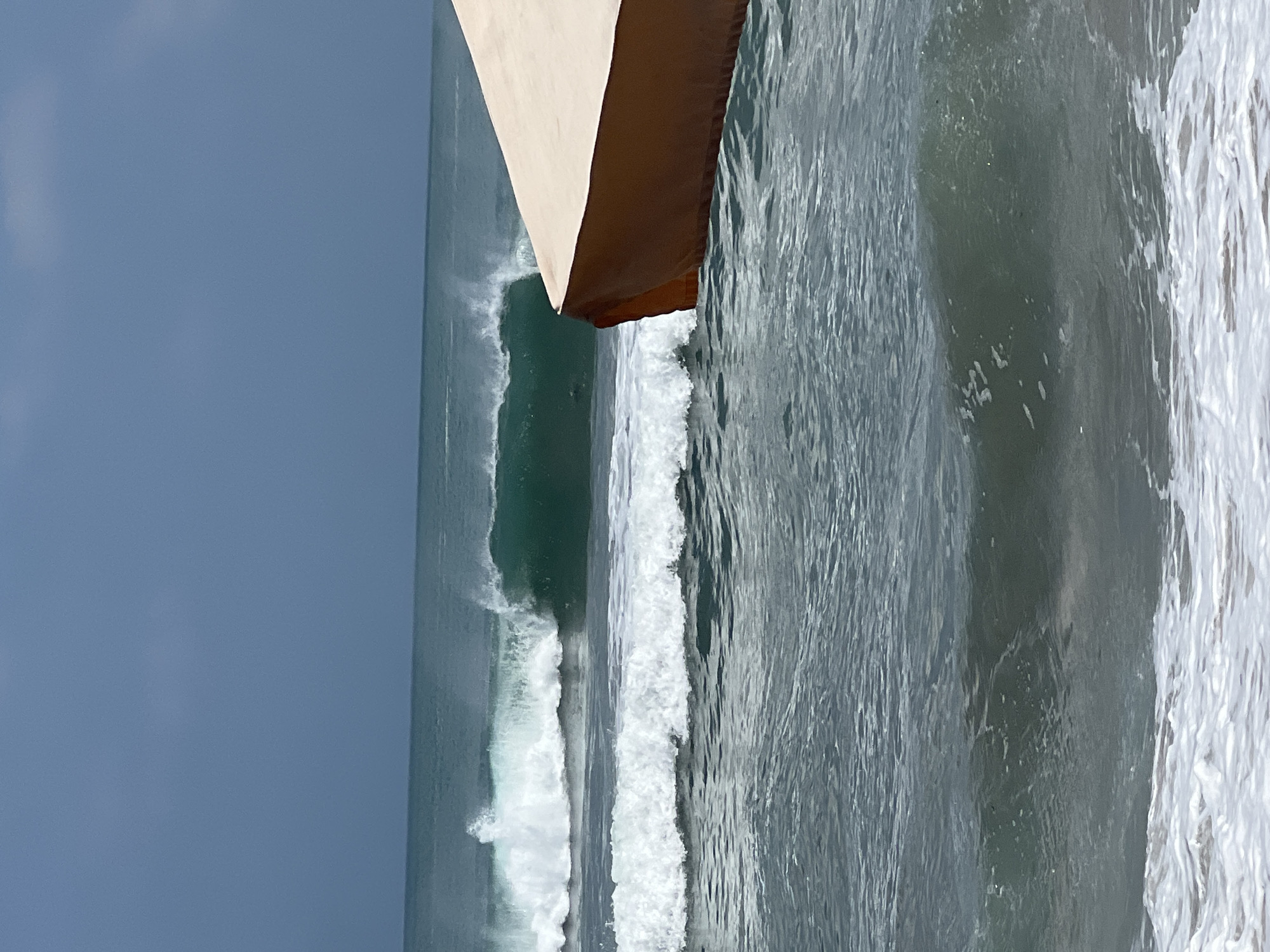 An overcast day at Kuta Beach, Indonesia, showcases a series of breaking waves. The ocean's surface is a mix of deep teal and lighter green hues, with white foam cresting the waves. In the foreground, the closest wave is gently rolling onto the shore, its white foam spread across the surface. In the mid-ground, larger waves are actively breaking, their emerald faces illuminated against the darker ocean. The background is dominated by a vast, muted blue sky, with the horizon line of the ocean barely distinguishable. To the right side of the frame, a large, tan-colored beach umbrella partially obscures the view of the sea, its brown underside visible. No people are visible in the scene.